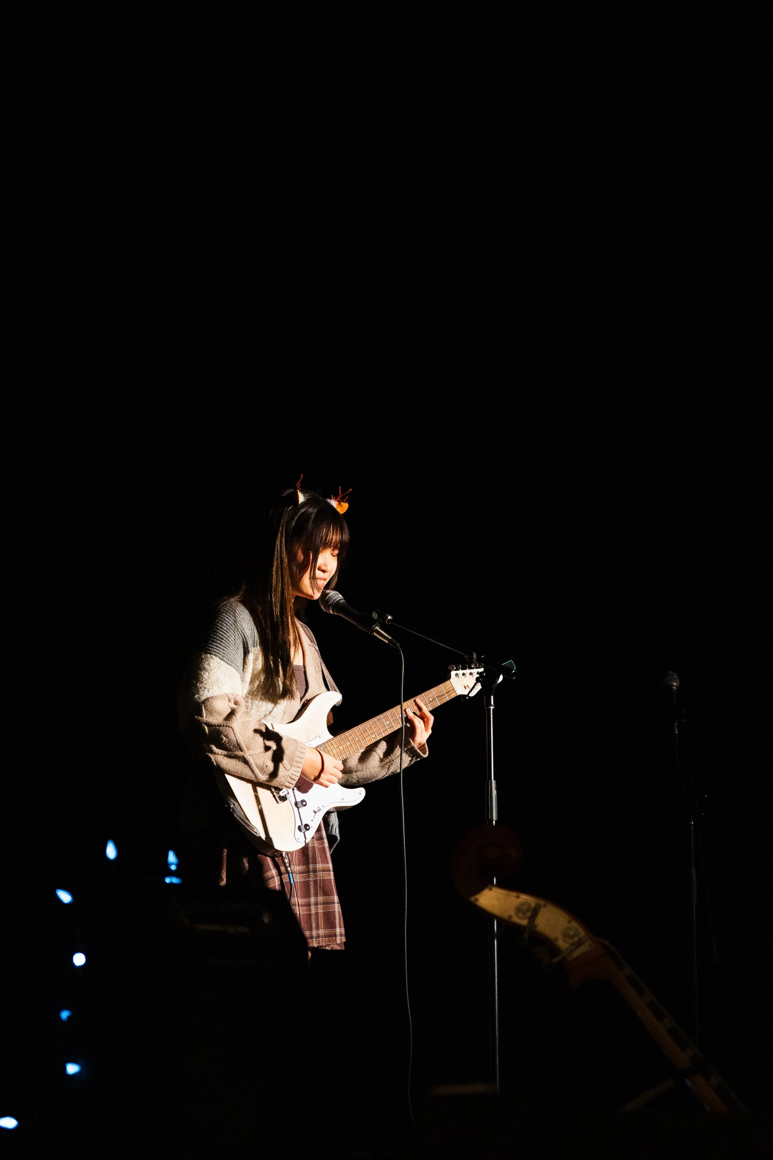 Young woman with long hair wearing a patterned jacket, playing electric guitar and singing into a microphone on a dark stage.