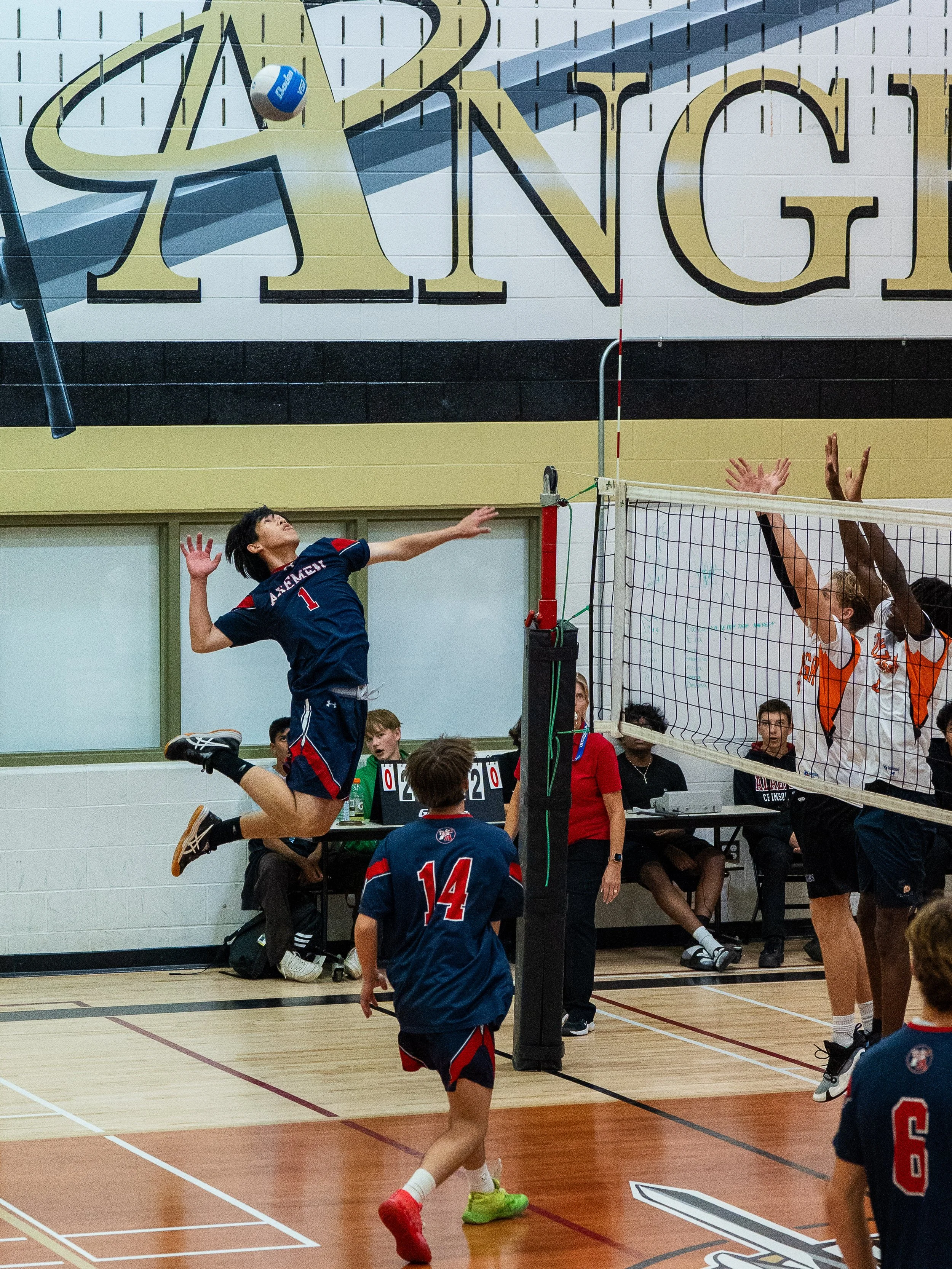 A volleyball game in progress with a player in mid-air hitting the ball over the net, while players on the opposing side jump to block. Several players and spectators are visible in the background.