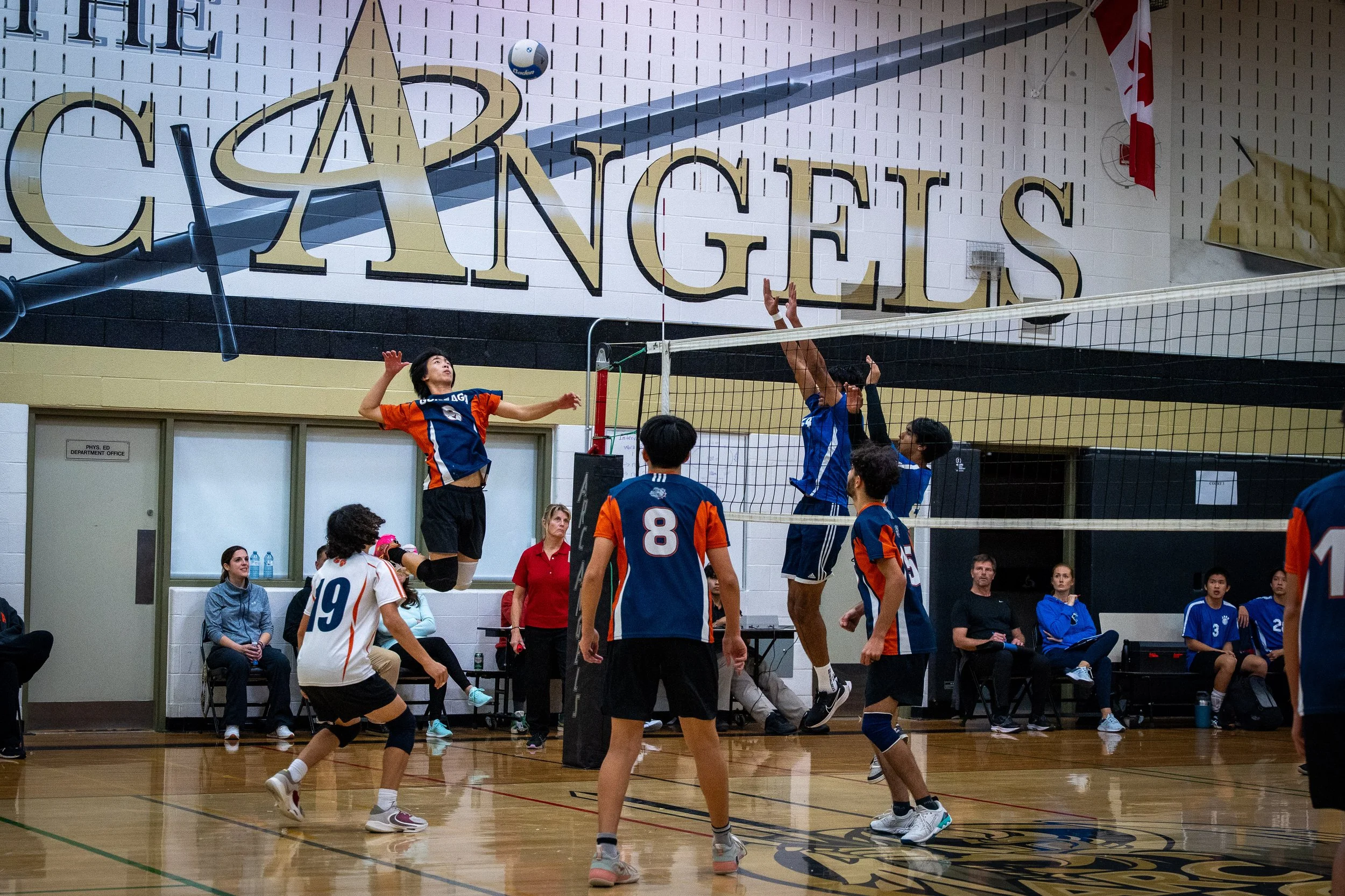 A volleyball game in progress in a gymnasium with players in blue and orange uniforms. One player is jumping to spike the ball while two players on the opposing team attempt to block it. Several spectators and coaches are seated and watching.