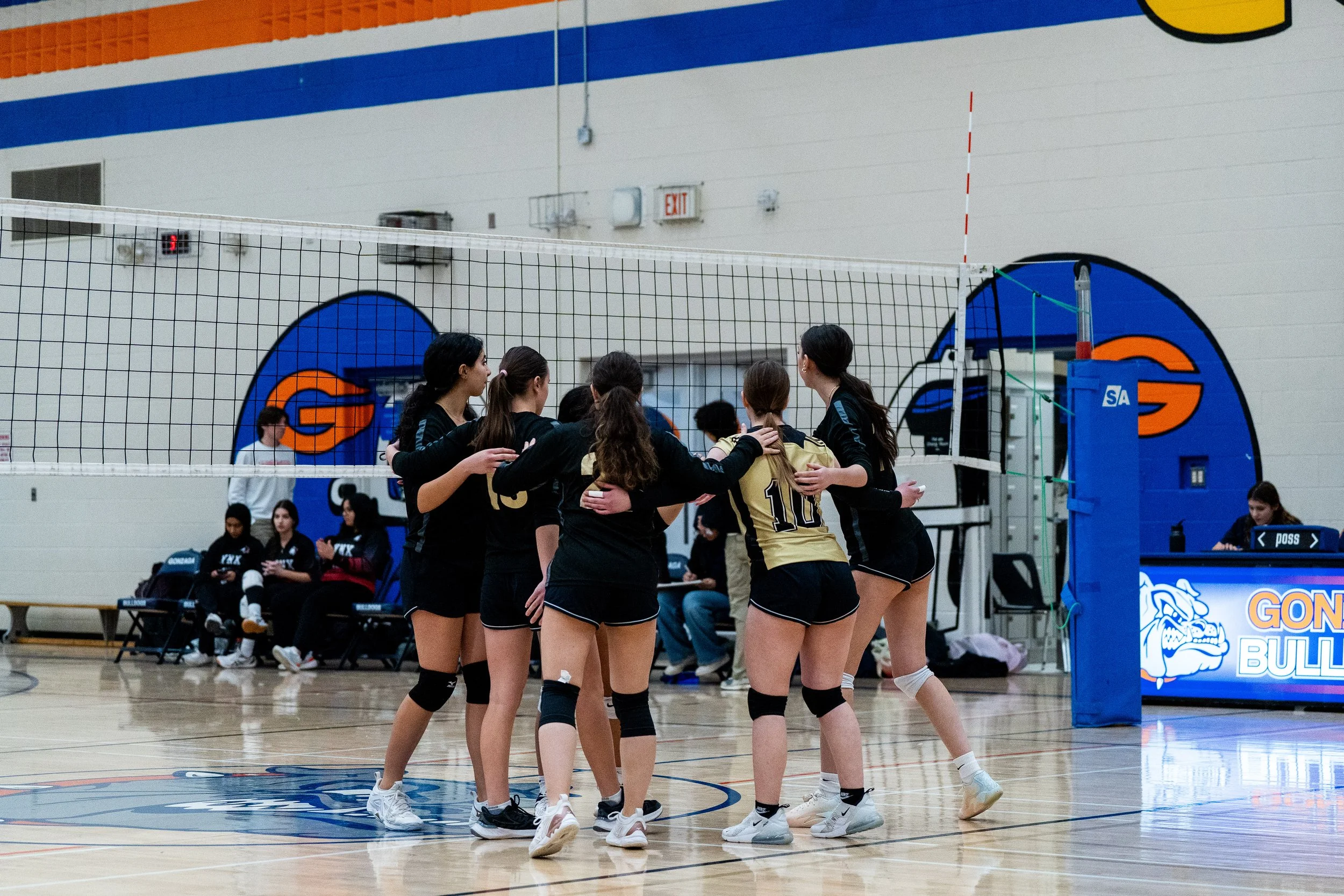 A group of female volleyball players in black and gold uniforms celebrating on an indoor court.