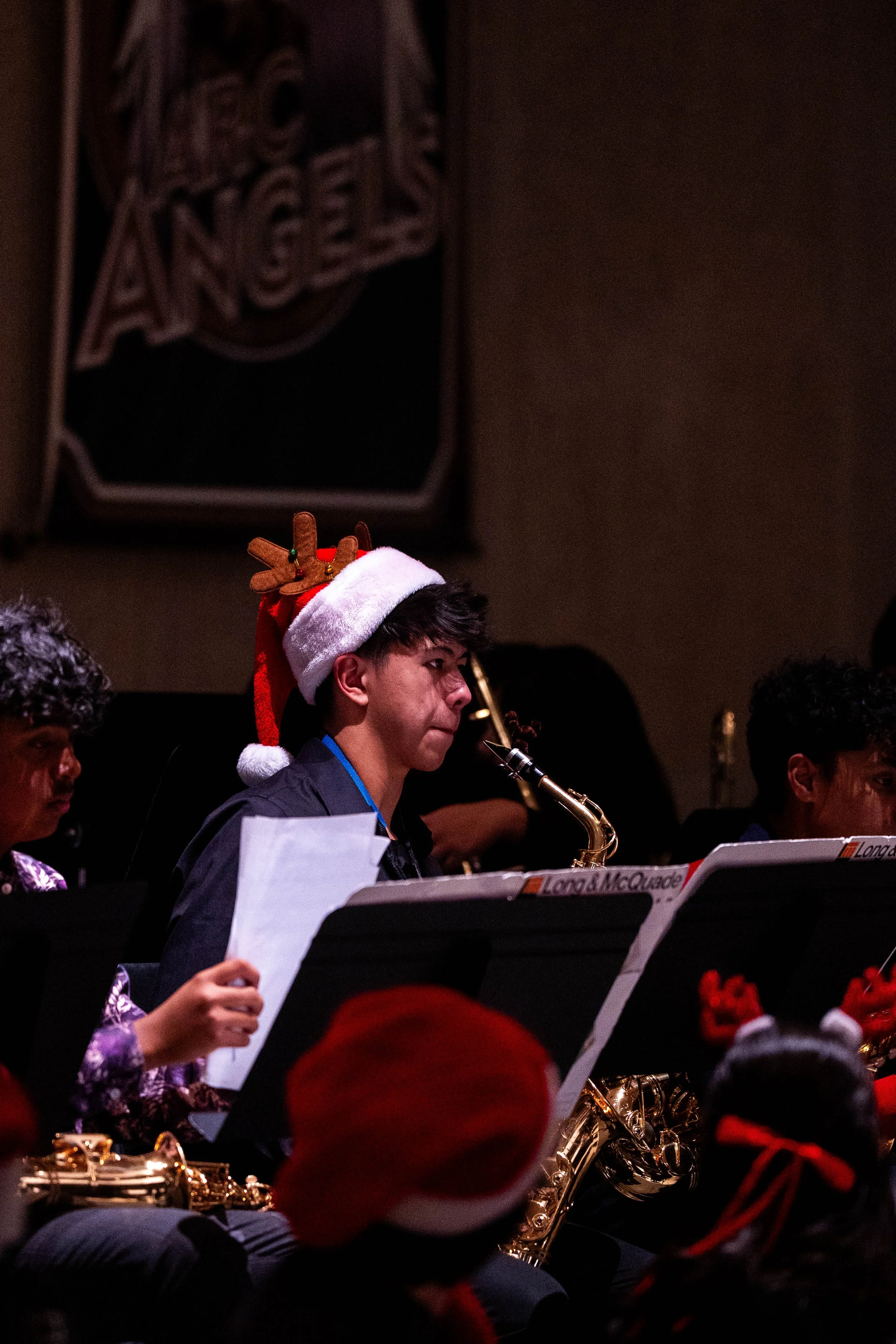 Young man playing saxophone in a Christmas-themed concert, wearing a Santa hat with reindeer antlers, surrounded by other musicians, some wearing Christmas accessories.