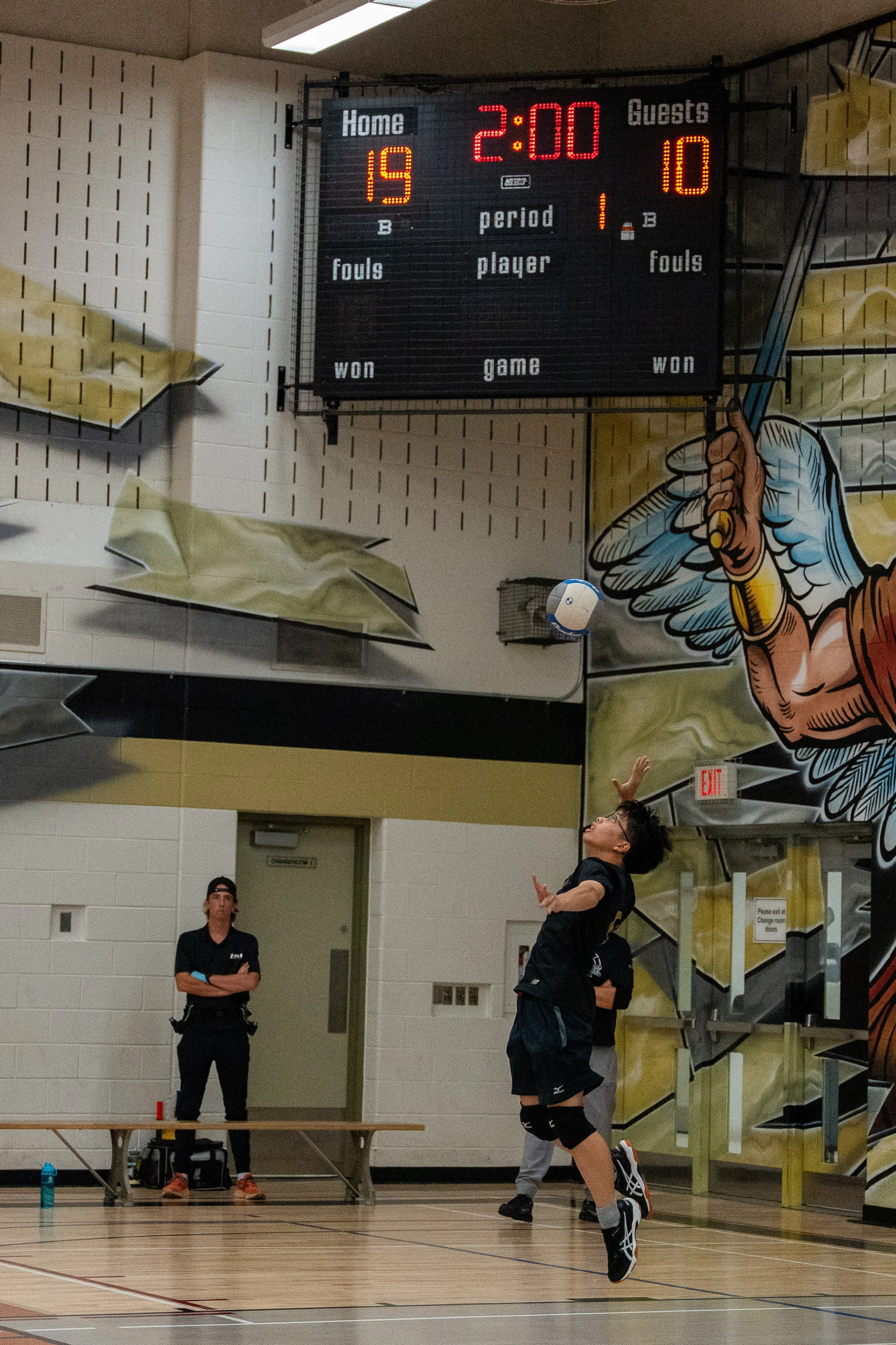 A volleyball game in progress with a player jumping to hit the ball as a referee watches. The scoreboard shows Home team 19, Guests 10, with 2 minutes remaining in the first period.