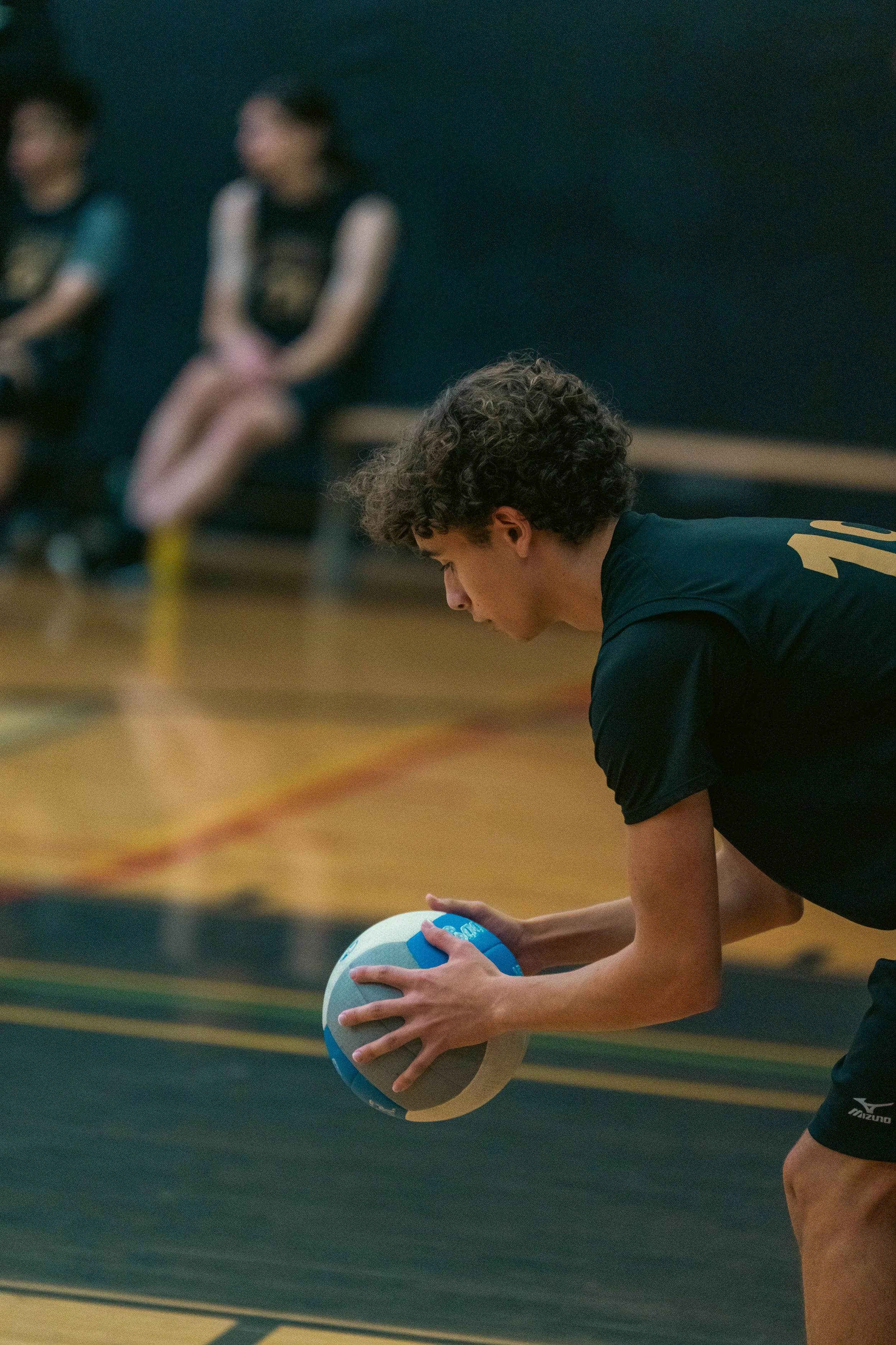 A young male volleyball player in black sportswear preparing to serve or pass the ball on an indoor court. Two other players are sitting on a bench in the background.