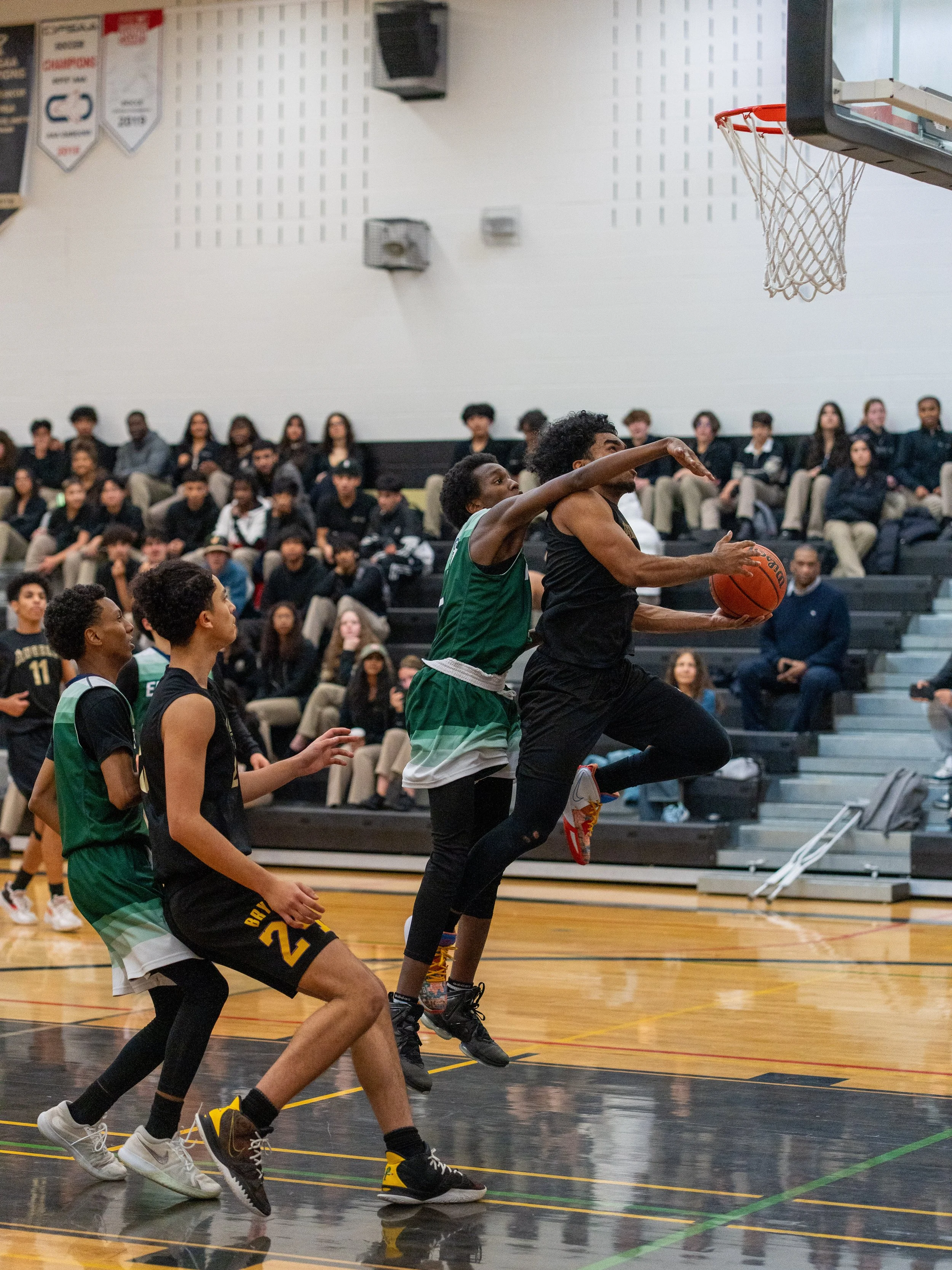 A basketball game in progress with a player in black jumping towards the hoop with the ball, while a player in green tries to block him. Other players and spectators are seated in the background.