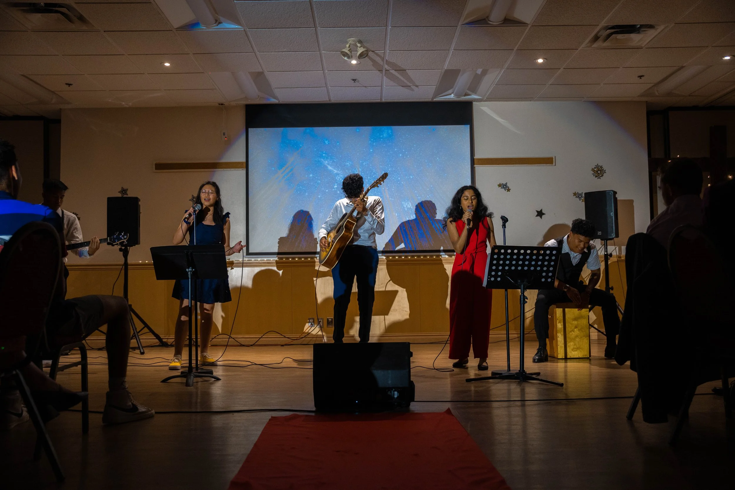 Group of five young musicians performing on stage with microphones, guitars, and percussion instrument in a decorated indoor venue, audience visible seated around the stage.