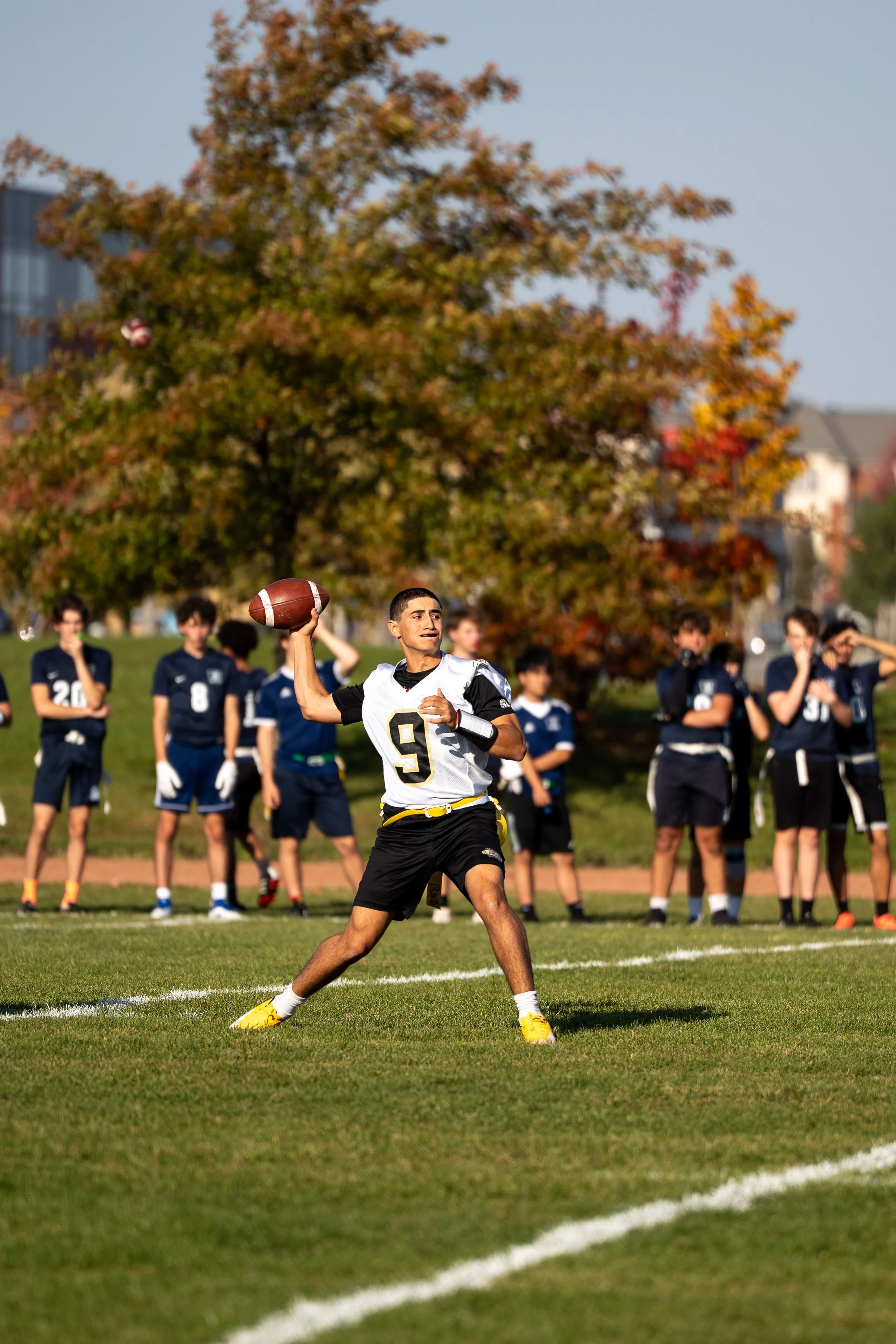 A young male football player in uniform prepares to throw a football on a field during game day. A group of teammates and coaches are in the background, along with colorful autumn trees.