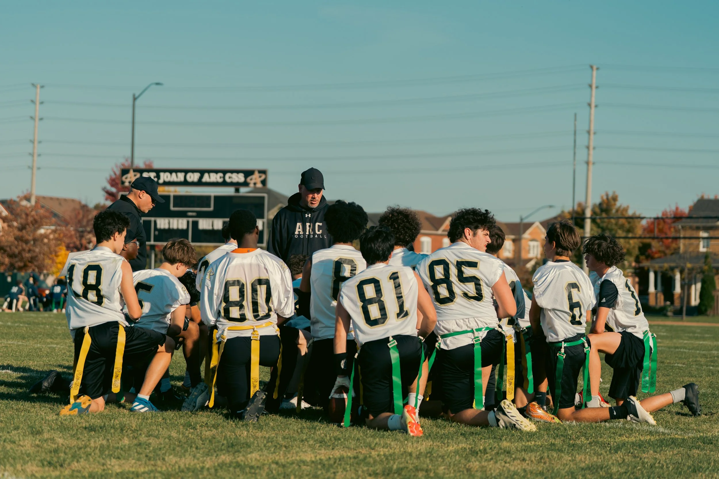 A youth football team kneeling in a huddle on a field with two coaches standing and talking to them, during a game or practice, with a scoreboard and houses in the background.