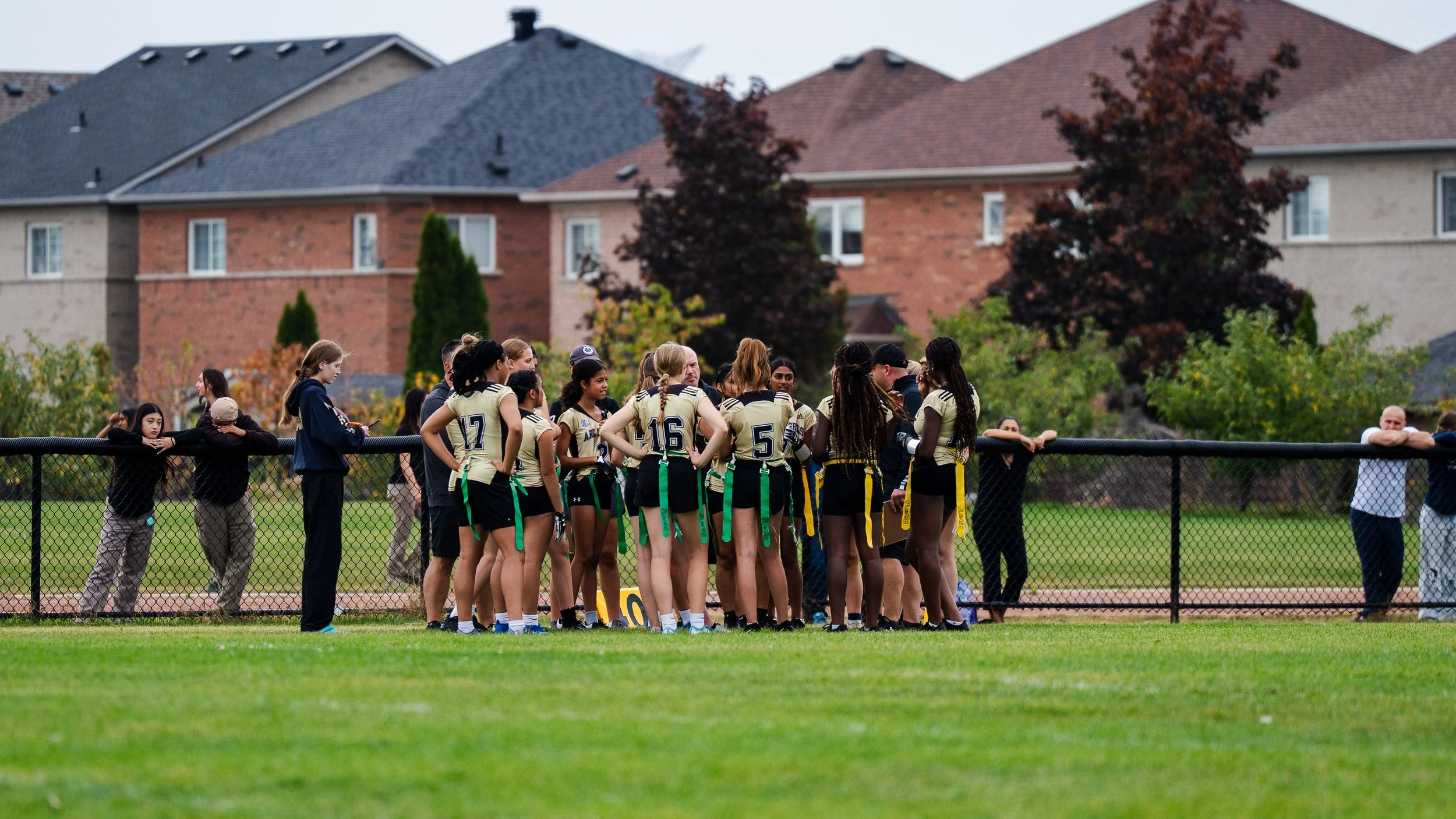 A group of young female soccer players and coaches gathered on a soccer field, listening attentively to a coach during a team meeting, with spectators standing behind a fence and suburban houses in the background.