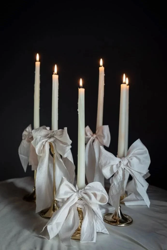 Five tall white candles in gold holders decorated with white satin bows, lit and placed on a white surface against a dark background.