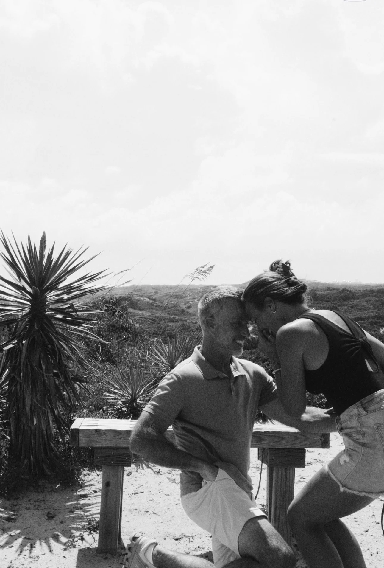 A black-and-white photo of a man kneeling with a woman leaning in close, touching foreheads and smiling, outdoors on a sunny day with desert plants and hills in the background.