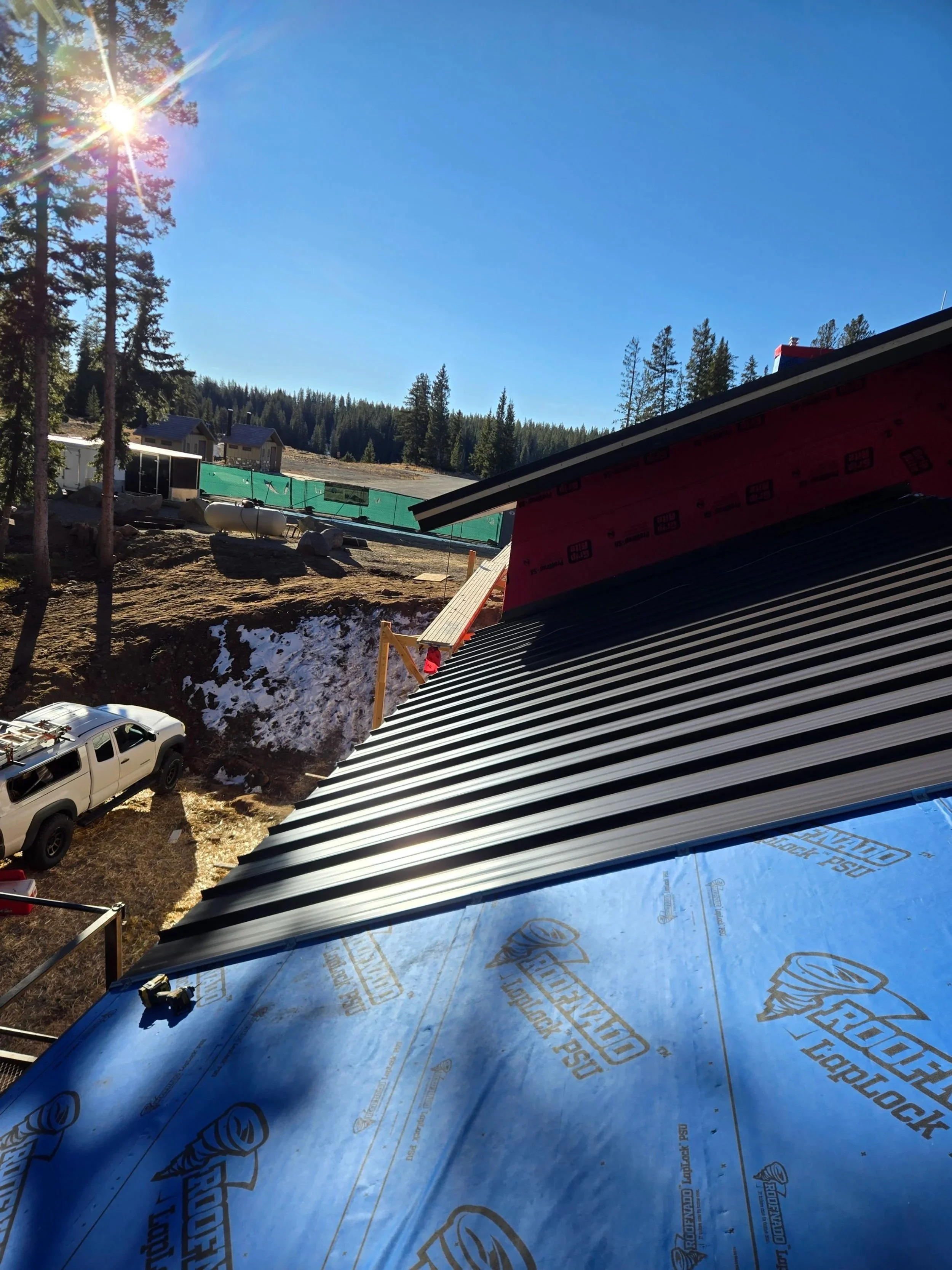 Construcción de un tejado con láminas metálicas en un área montañosa, rodeada de árboles y viviendas, con un cielo despejado y el sol brillando.