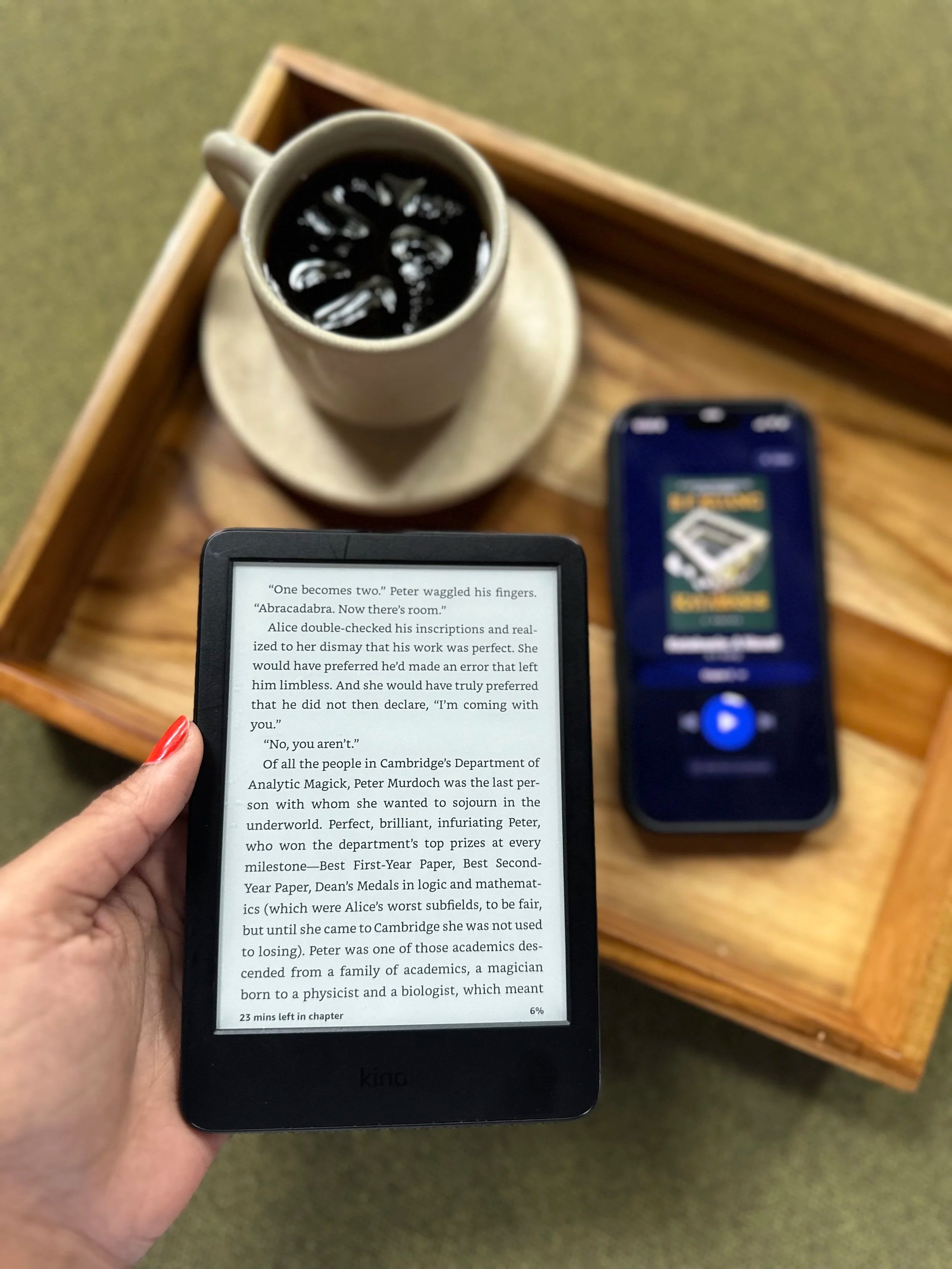 A person holding a Kindle e-reader displaying a page of text, with a wooden tray holding a coffee cup filled with dark coffee and an iPhone showing an audio player app, all placed on a table.