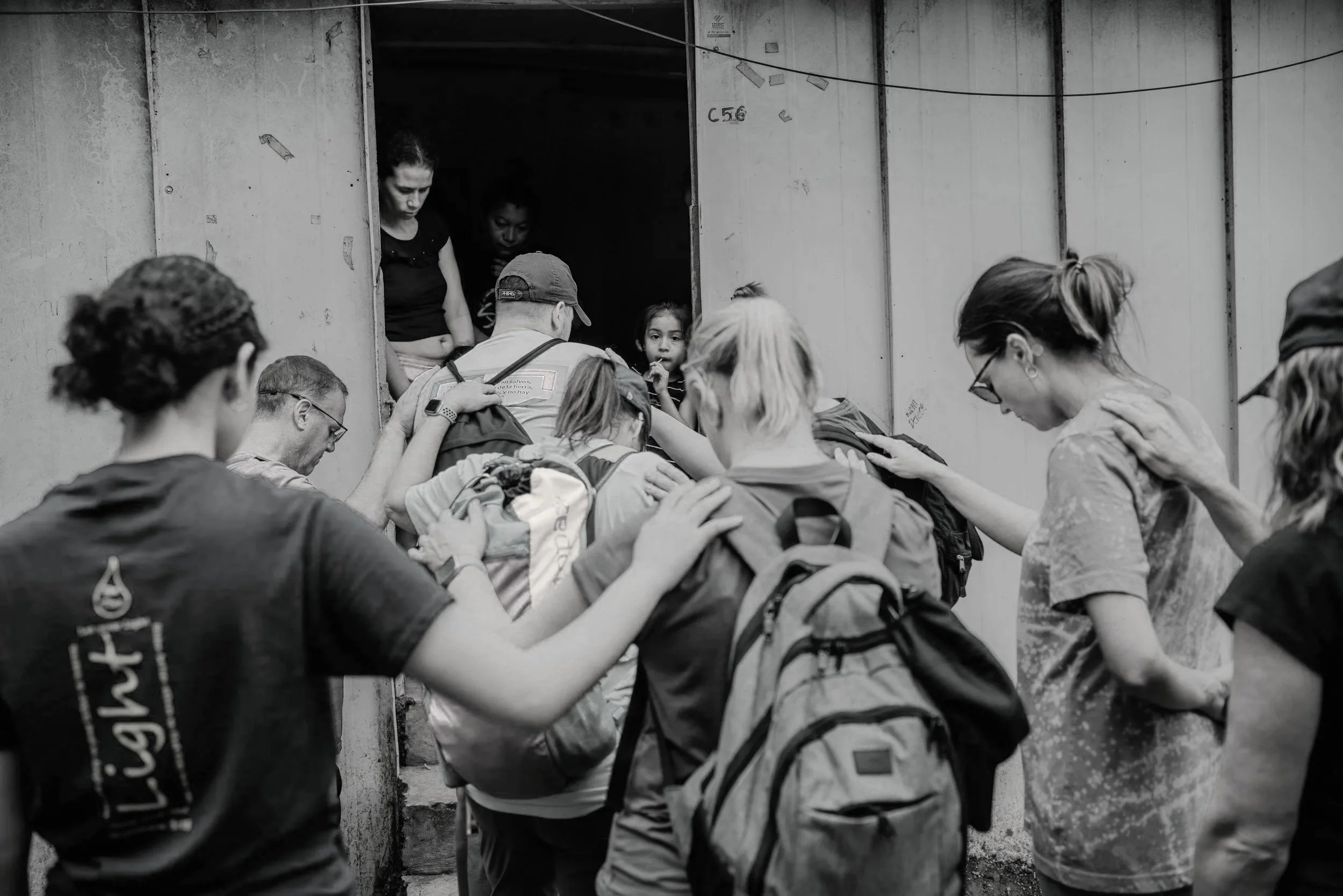 Group of children and adults praying or making a prayer circle outside a building with a door, some with backpacks, in black and white.