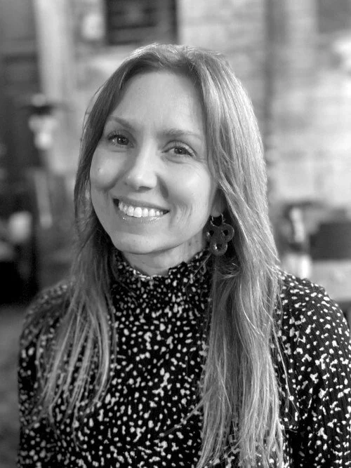 Black and white portrait of a smiling woman with long hair, wearing a patterned top and large earrings, in an indoor setting.