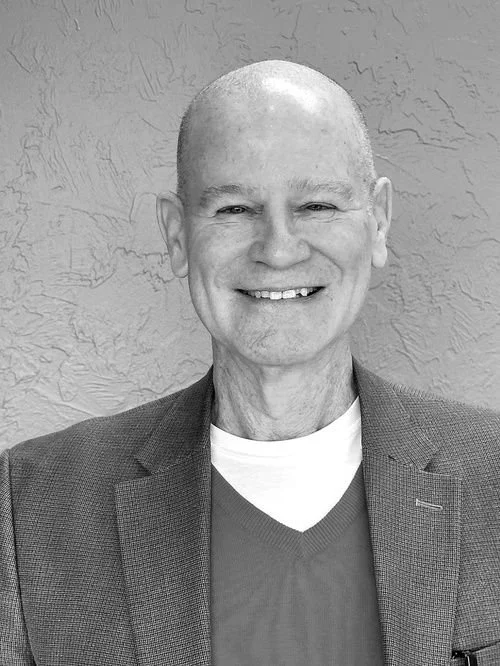 A smiling, bald man wearing a blazer and a t-shirt, standing against a textured wall background.
