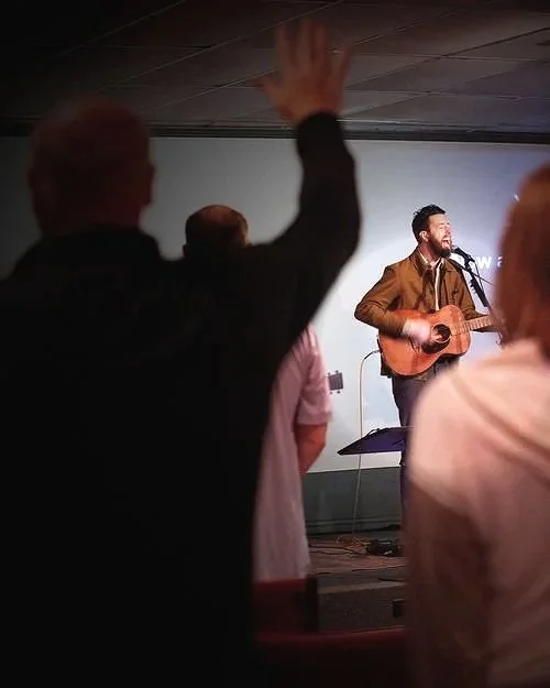 A musician with a guitar performing on stage in front of a small audience, some of whom are raising their hands.