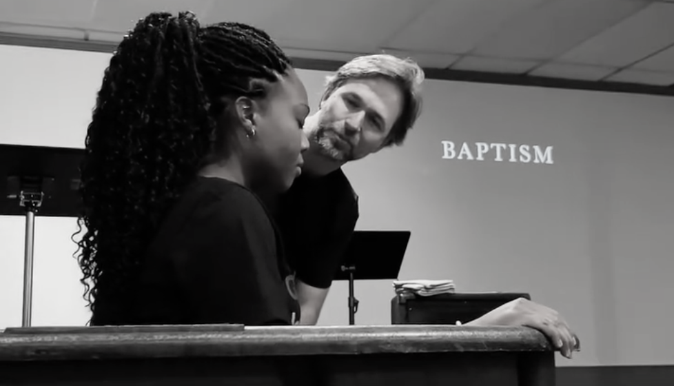 A woman and a man are having a conversation at a baptism ceremony, with a sign that reads 'BAPTISM' in the background.