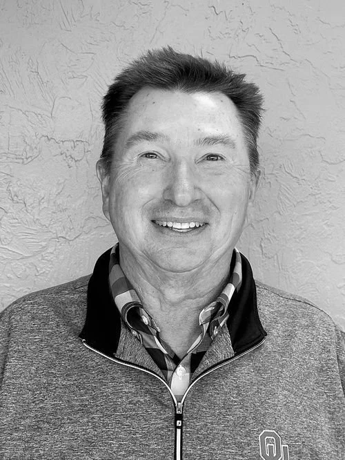 A smiling man with short dark hair, wearing a checkered shirt under a zip-up sweater with an 'Oklahoma' logo, standing against a textured wall.