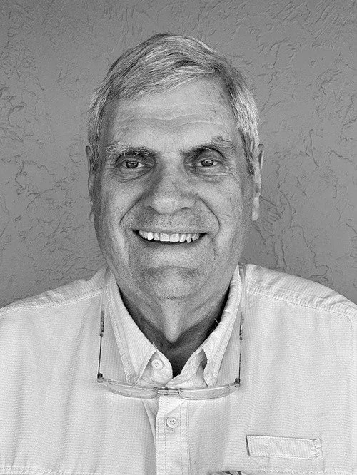 Black and white photo of smiling middle-aged man with glasses hanging from collar, short gray hair, dressed in a collared shirt, against textured wall background.