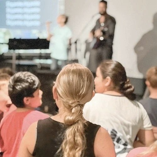 People attending a live performance or talk, listening to a woman speaking and a man standing nearby, with an audience in the foreground.