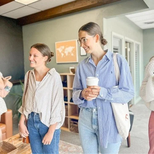 Two women smiling and talking in an indoor setting, one holding a coffee cup. The Orchard at Mission.