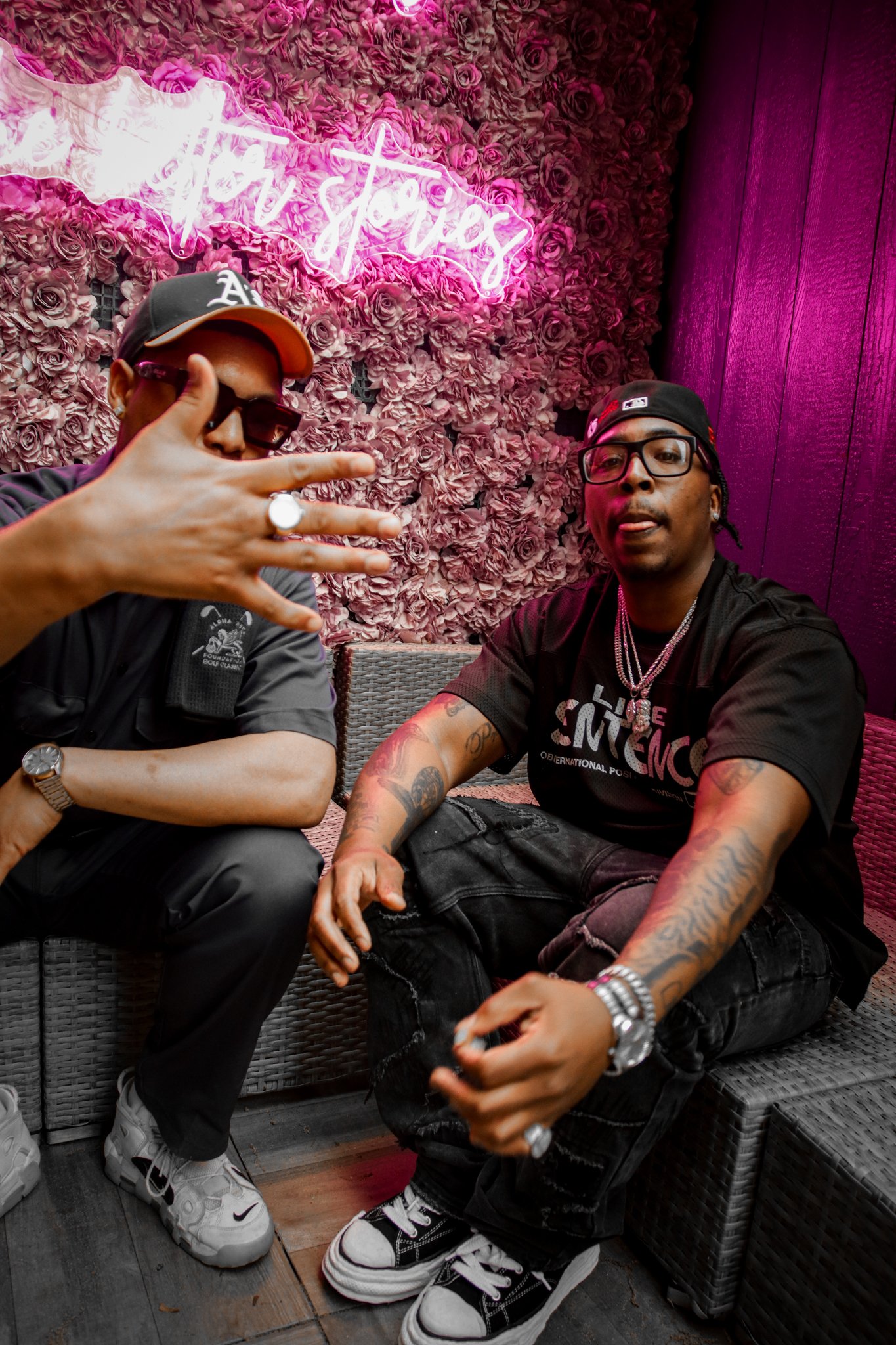 Two men sitting on rattan furniture in front of a pink rose wall with neon sign that reads 'Bar Stays'. One is wearing a black T-shirt, ripped black jeans, a black cap, and glasses, while the other is dressed in black pants, white sneakers, sunglasse