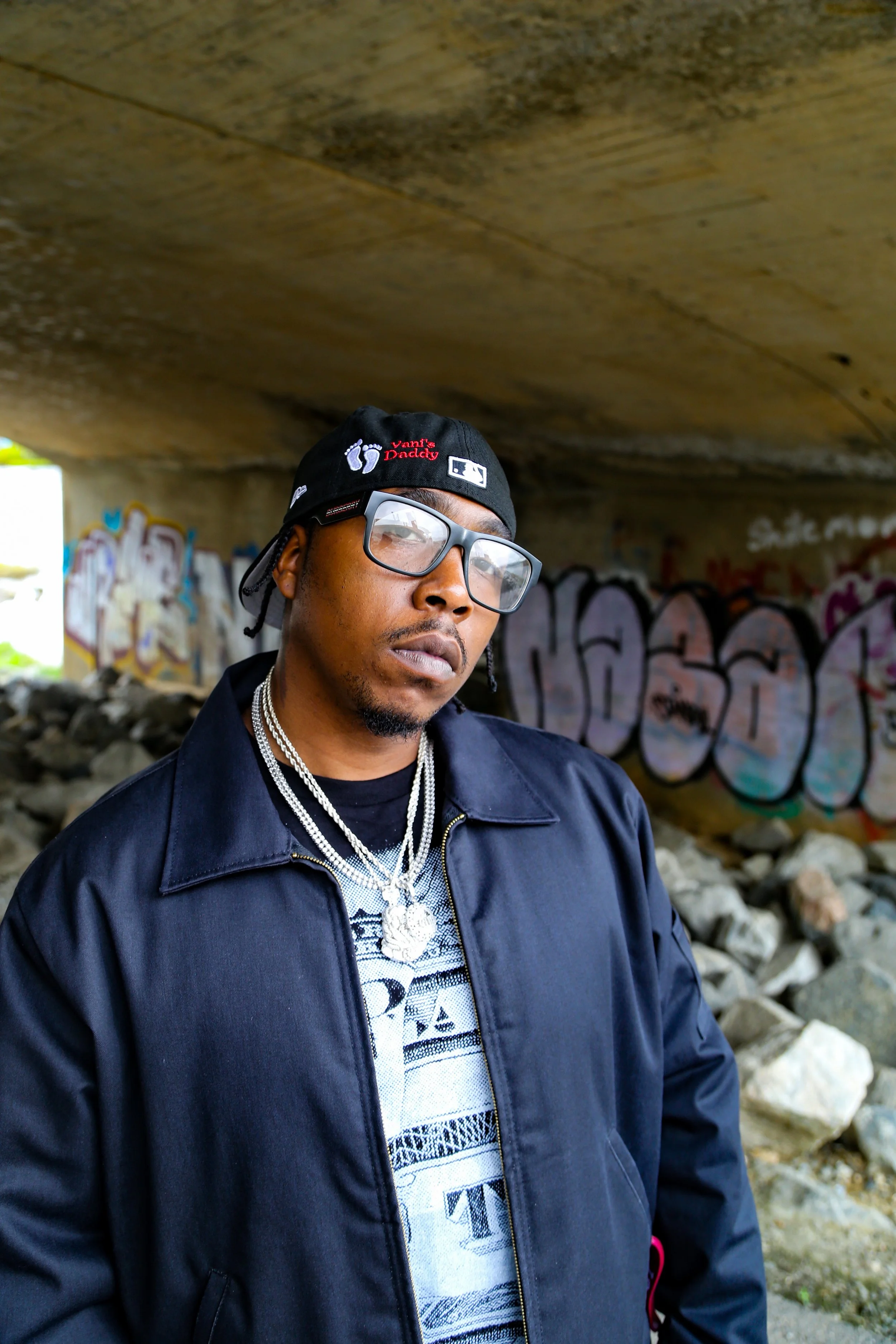 A man wearing glasses, a black jacket, and layered silver necklaces, standing under a graffiti-covered bridge with rocks and debris on the ground.