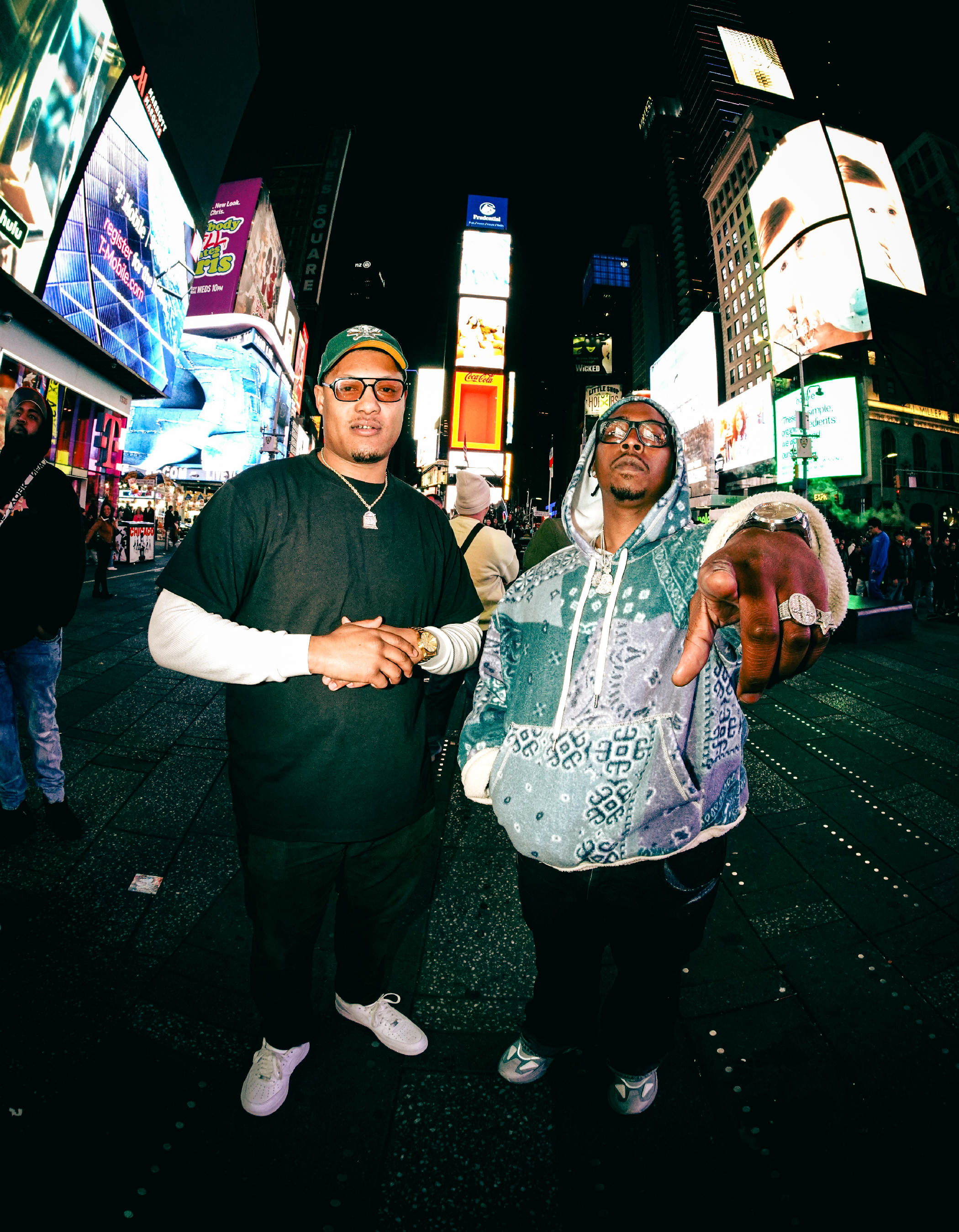 Two men standing in Times Square at night with bright digital billboards in the background. The man on the left is wearing glasses, a black and white long-sleeve shirt, and white sneakers. The man on the right is wearing sunglasses, a patterned hoodi