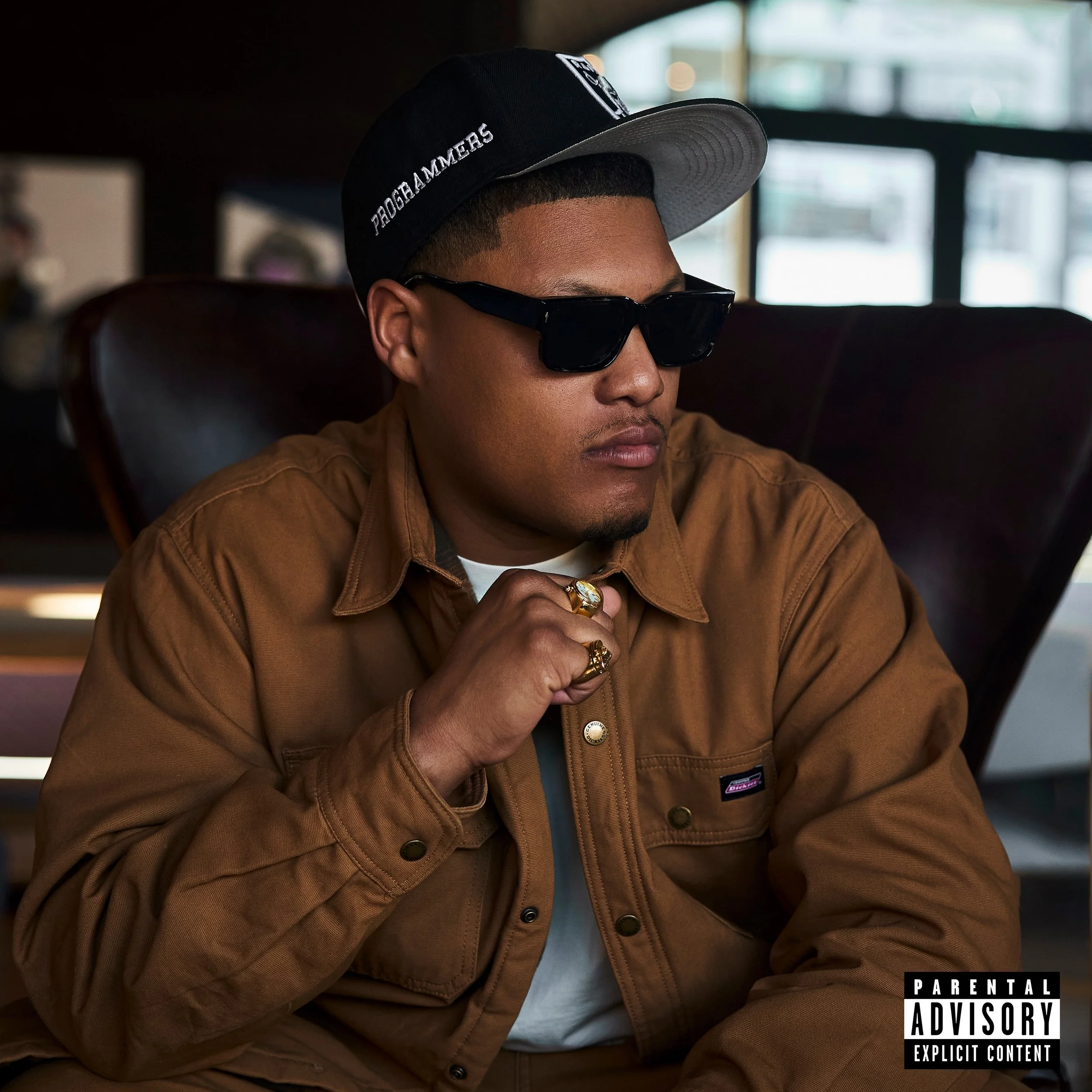 A young man wearing a black cap with white text, black sunglasses, and a brown jacket sitting in a vehicle with a blurred background of windows.