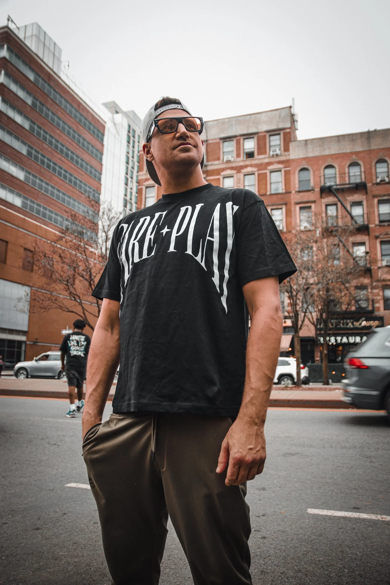 A man in sunglasses and a black cap worn backwards stands in front of a city street with tall buildings, a few trees, and passing cars. He is wearing a black T-shirt with the words 'CREATE + PLAY' printed on it and beige pants.