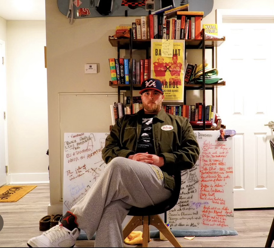 A man sits on a stool in front of a bookshelf filled with books and posters, with whiteboards filled with handwritten notes behind him, in a room with a door and a window.