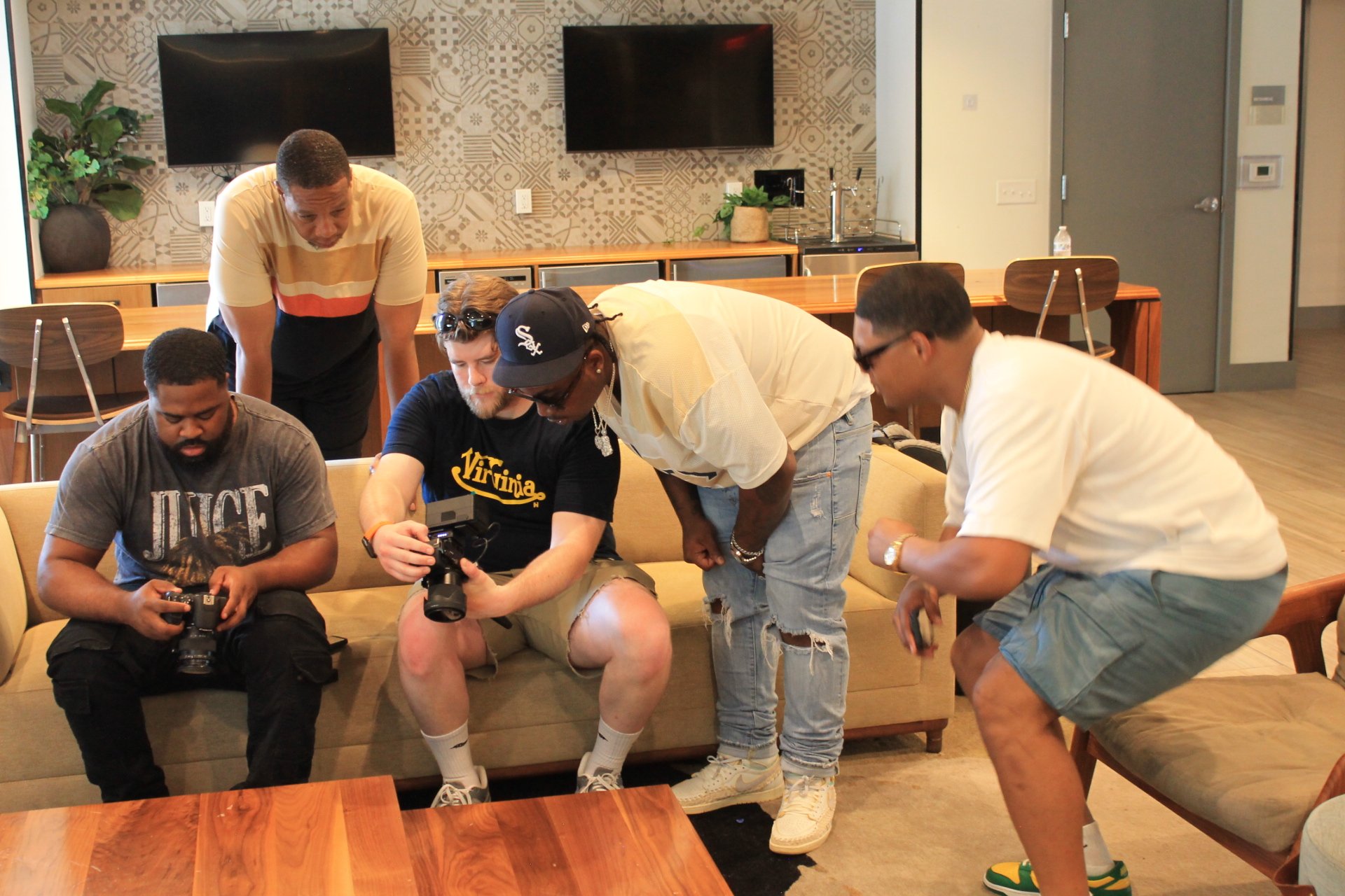 Group of five men gathered around a camera, looking at its screen, in a modern room with wall-mounted TVs and a wooden table.