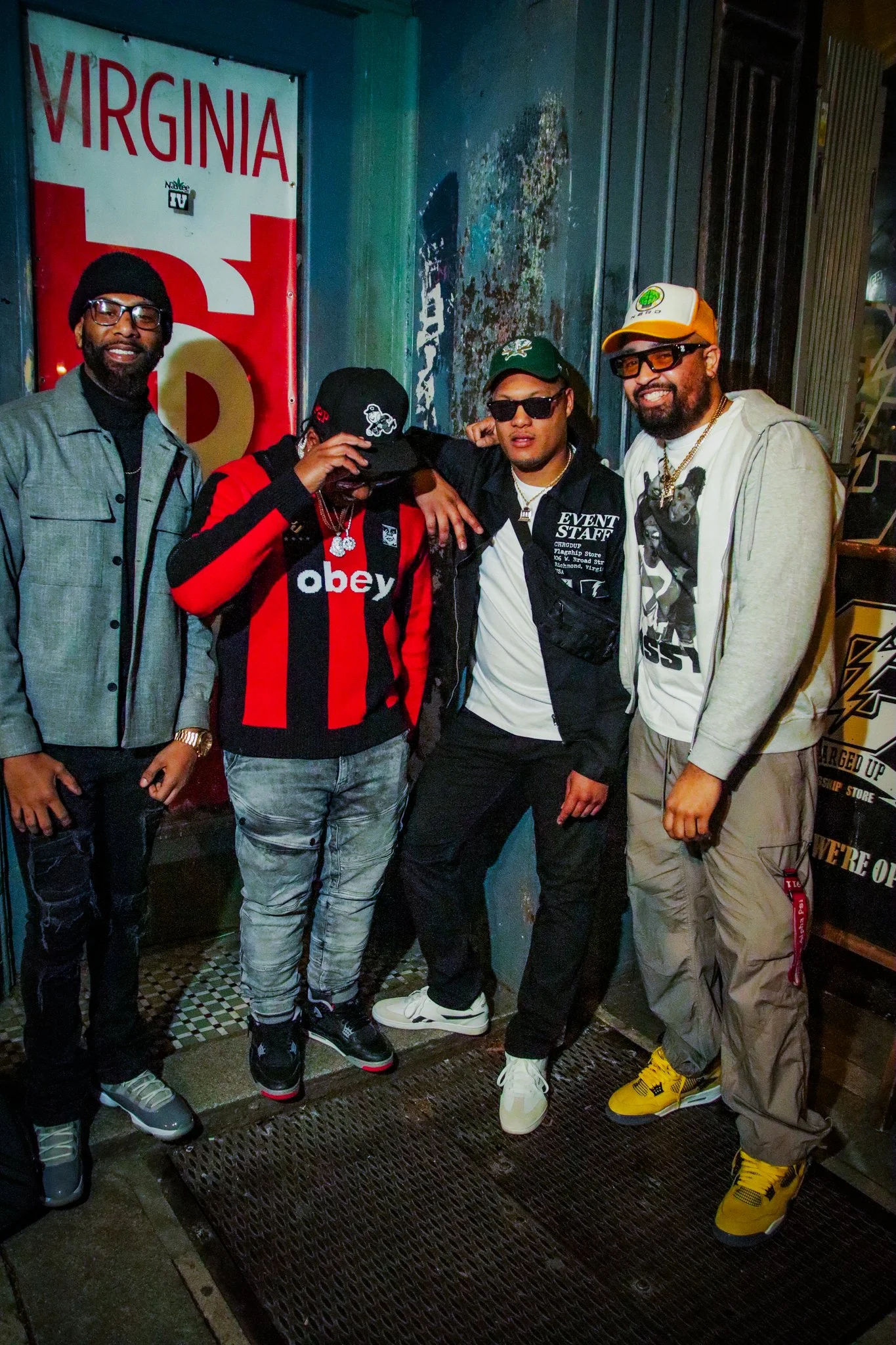 Four men standing together outside a building at night, with a Virginia license plate in the background.