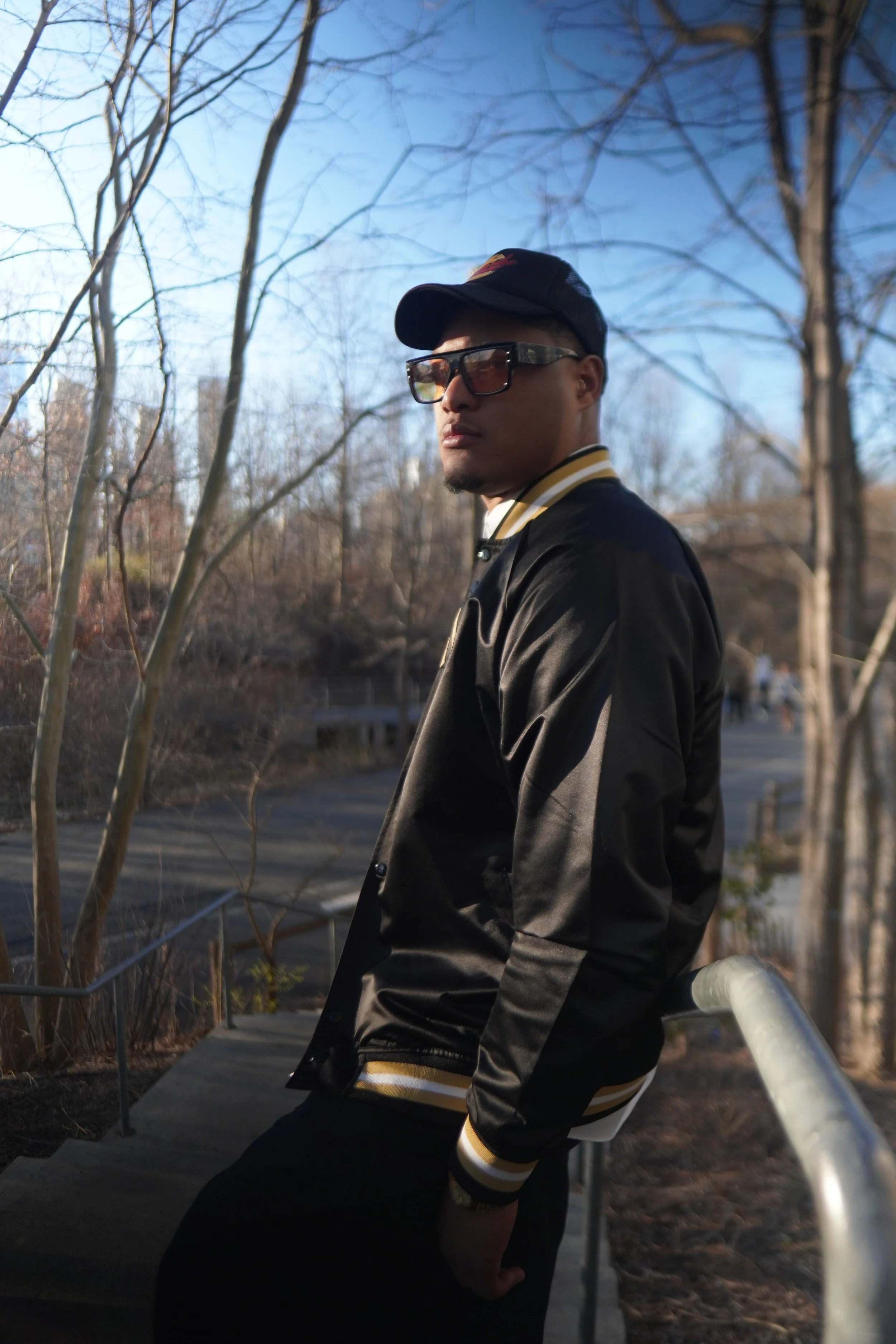 A man wearing sunglasses, a black cap, and a black jacket with yellow and white striped cuffs leaning against a railing outdoors with leafless trees in the background.