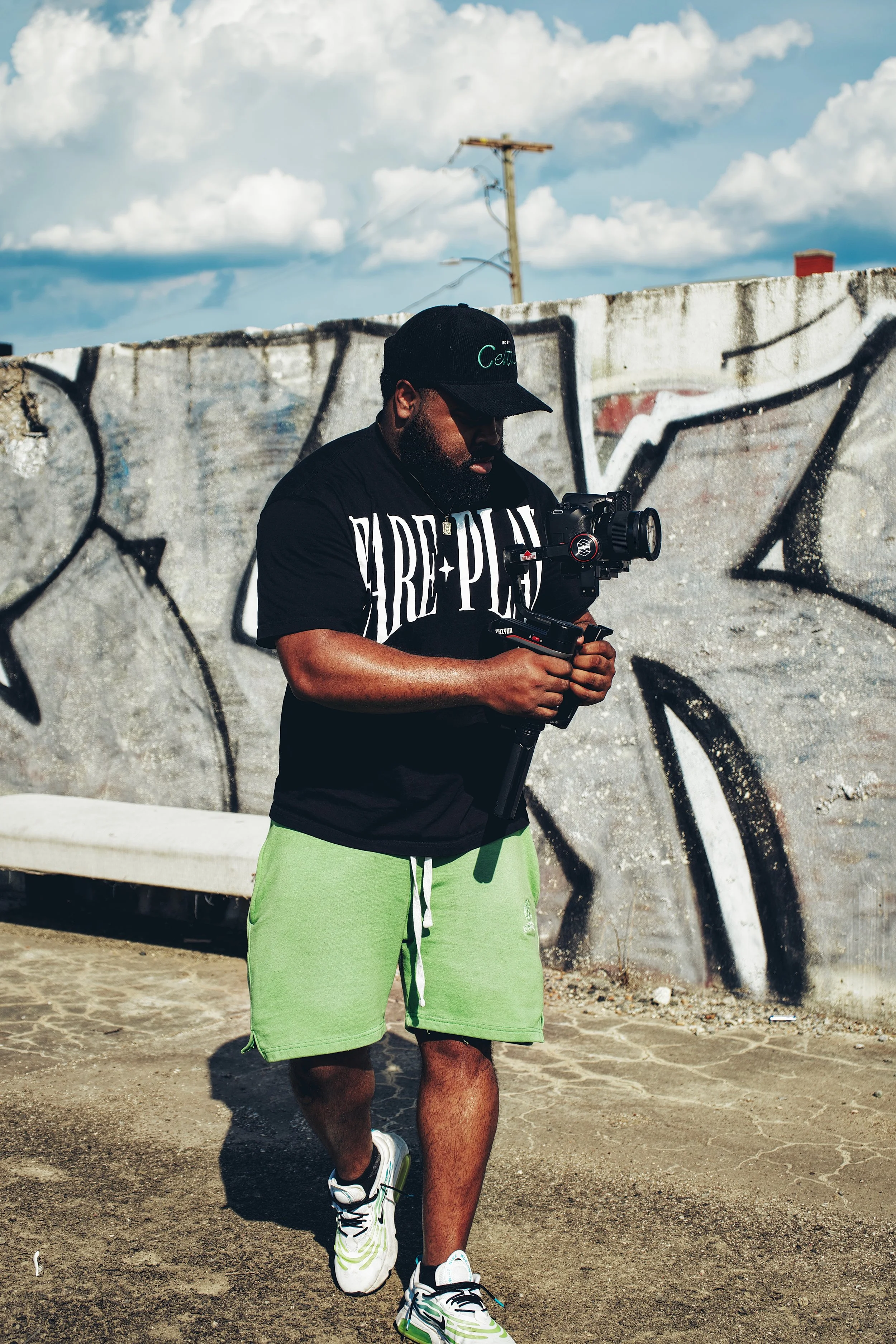 A man wearing a black cap, black T-shirt, green shorts, and white sneakers, standing outdoors near a graffiti-covered wall, holding a camera mounted on a stabilizer.