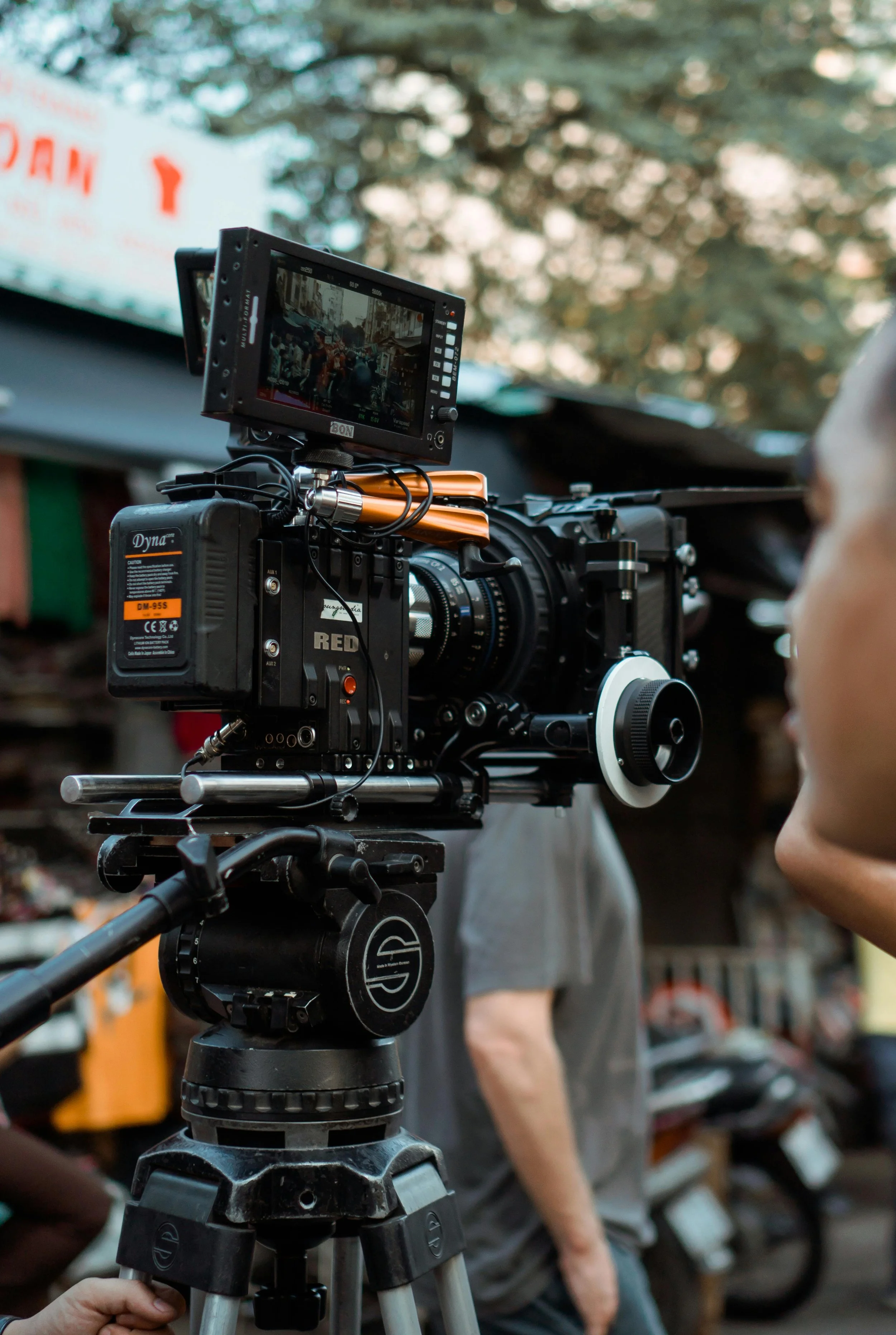 Close-up of a professional camera on a tripod, with a monitor attached, on a film set outdoors. Blurred background shows trees and people.