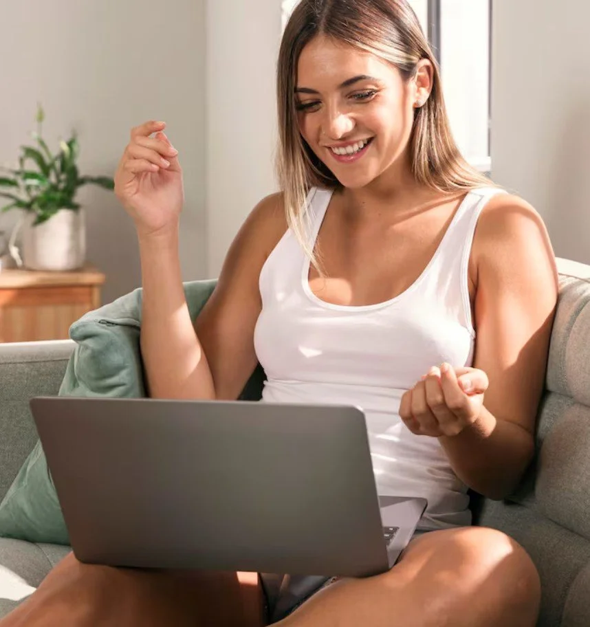 A woman sitting on a couch, smiling and celebrating while looking at her laptop in a well-lit living room.