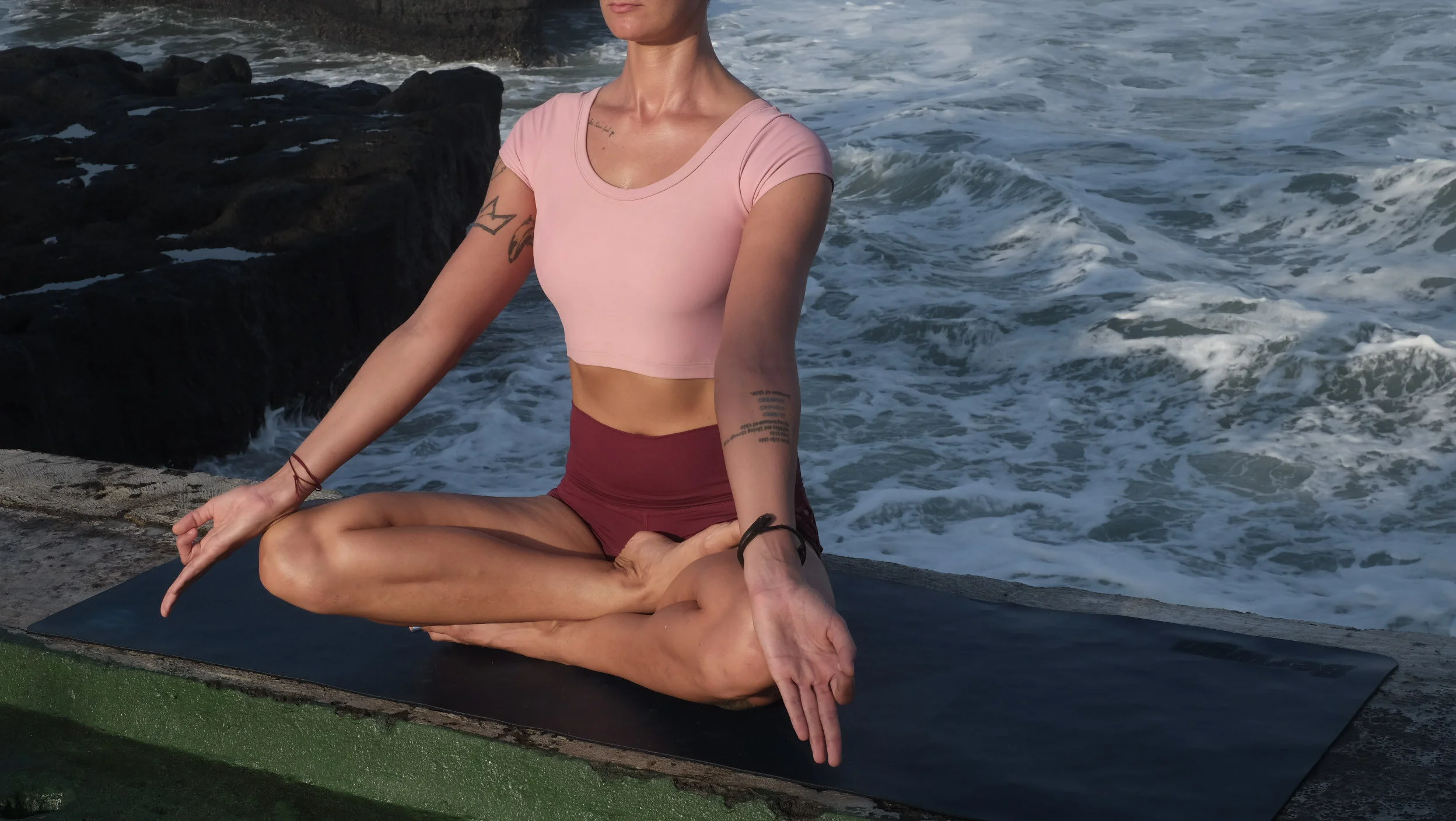 A woman practicing yoga in a seated lotus position on a mat near the ocean, with rocks and waves in the background.
