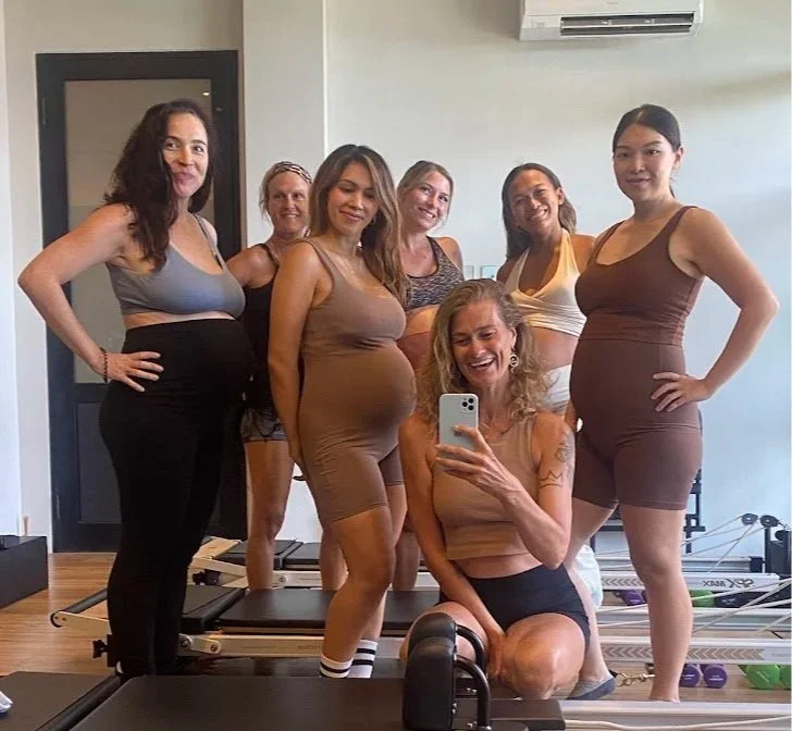 Group of seven women in workout clothes taking a selfie in a gym mirror, smiling.