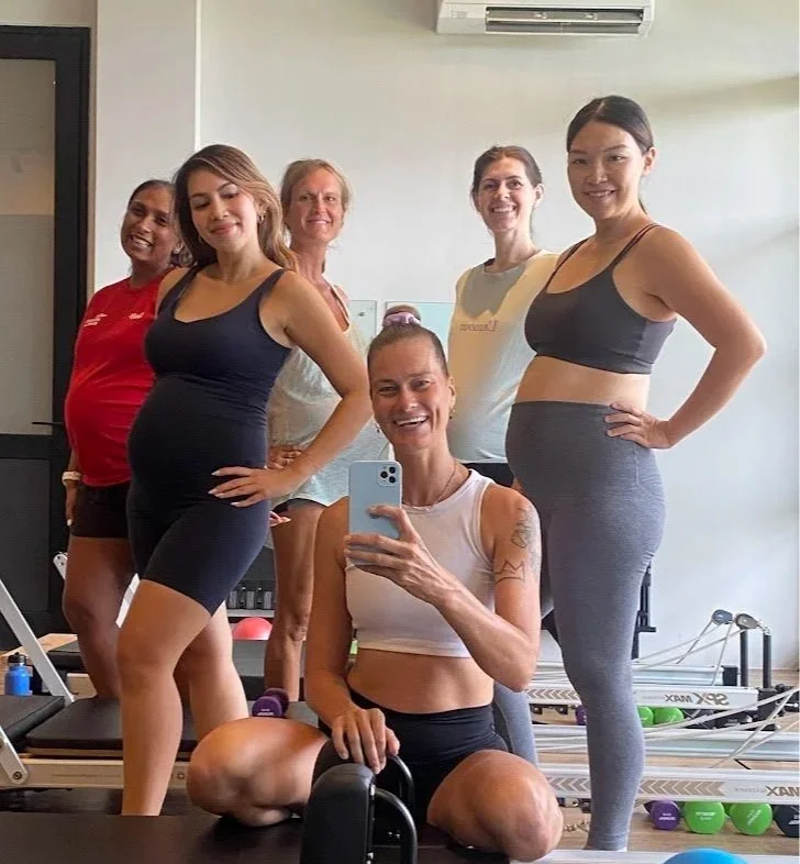 Group of five women posing for a selfie at a gym or fitness studio, with exercise equipment in the background.