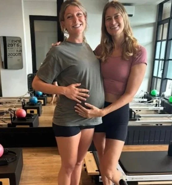 Two pregnant women smiling and holding their bellies at a Pilates studio with exercise equipment in the background.