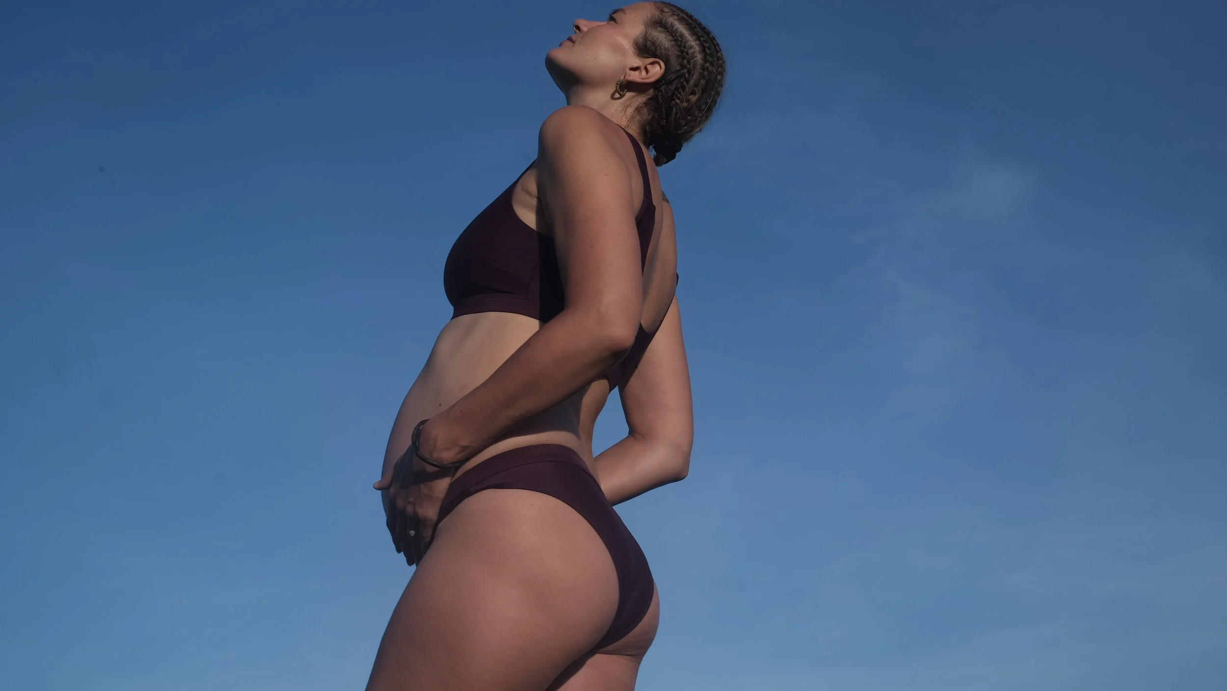 A woman in burgundy lingerie standing outdoors against a blue sky, with her eyes closed and head tilted back.