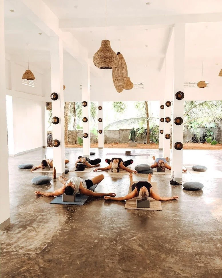 Group of people practicing yoga in a bright, spacious studio with large windows and tropical plants outside.