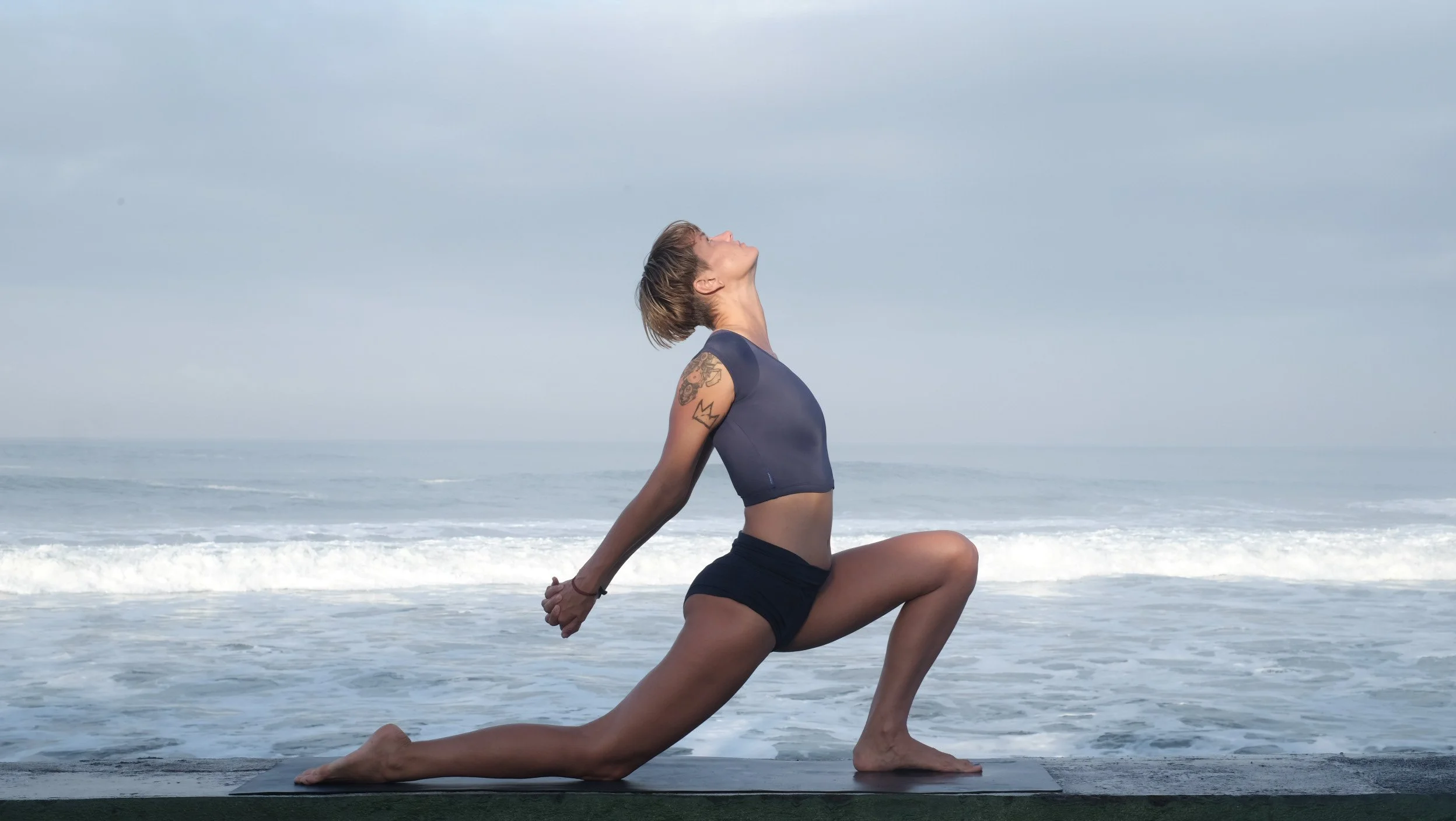 Woman practicing yoga on the beach during cloudy weather, in a low lunge pose with her head tilted back.