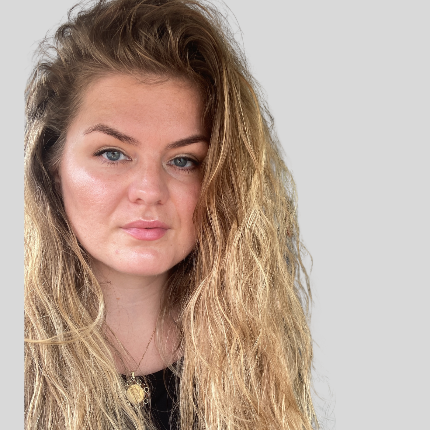 Close-up of a young woman with long wavy blonde hair, blue eyes, and a fair complexion, wearing a gold pendant necklace and a black top, against a plain light grey background.