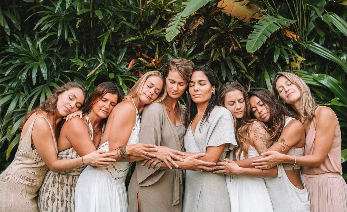 A group of eight women standing close together with their eyes closed, hugging and touching each other's arms, in front of lush green foliage.