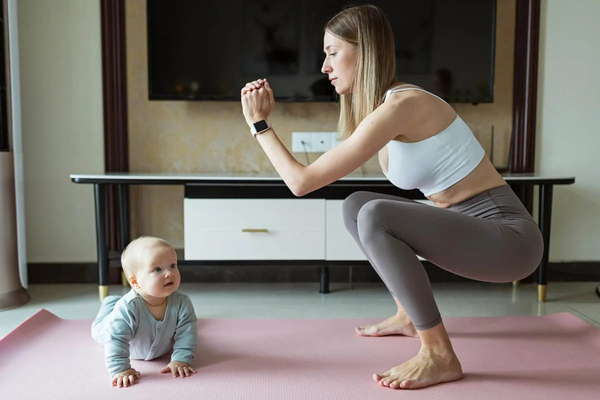 A woman in workout clothes is doing a squat on a pink yoga mat while a baby looks up at her from the ground.
