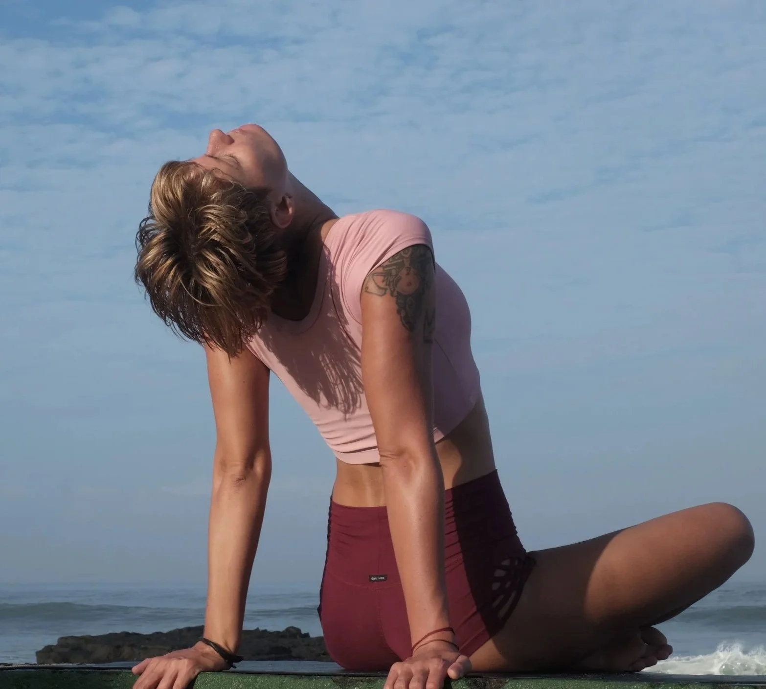 A woman practicing yoga outdoors near the ocean, performing a backbend stretch on a yoga mat.