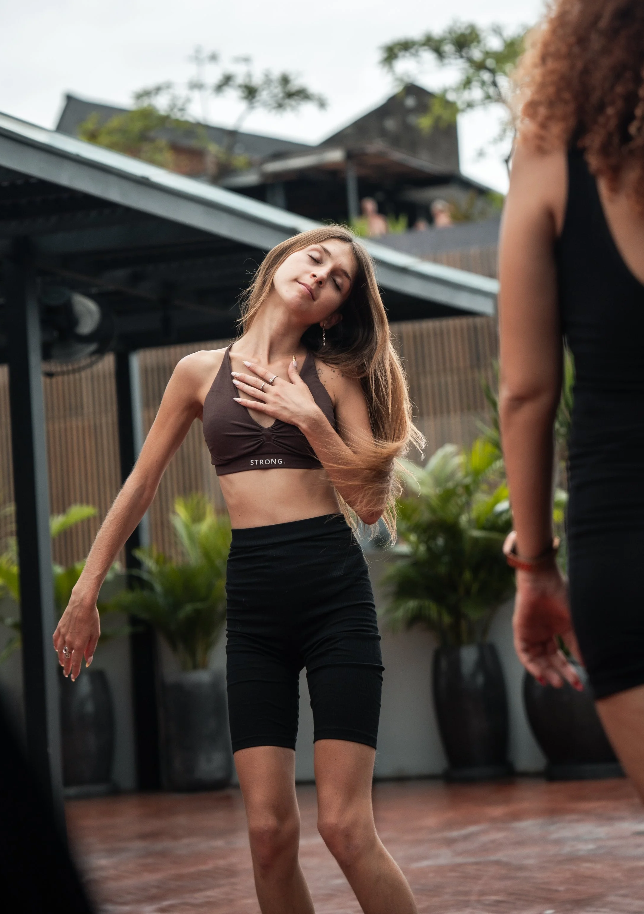 A woman with long hair wearing a black sports bra and black shorts dancing outdoors with her hand on her chest, eyes closed, and a relaxed expression.