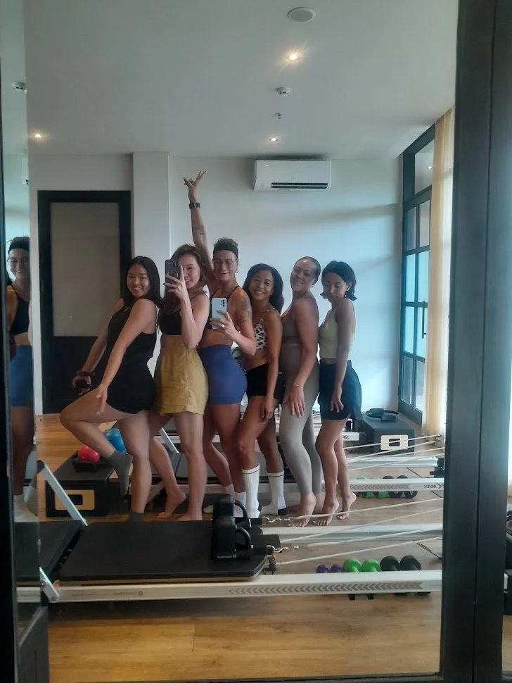Group of six women taking a mirror selfie in a fitness studio, with exercise equipment and colorful dumbbells on the floor.